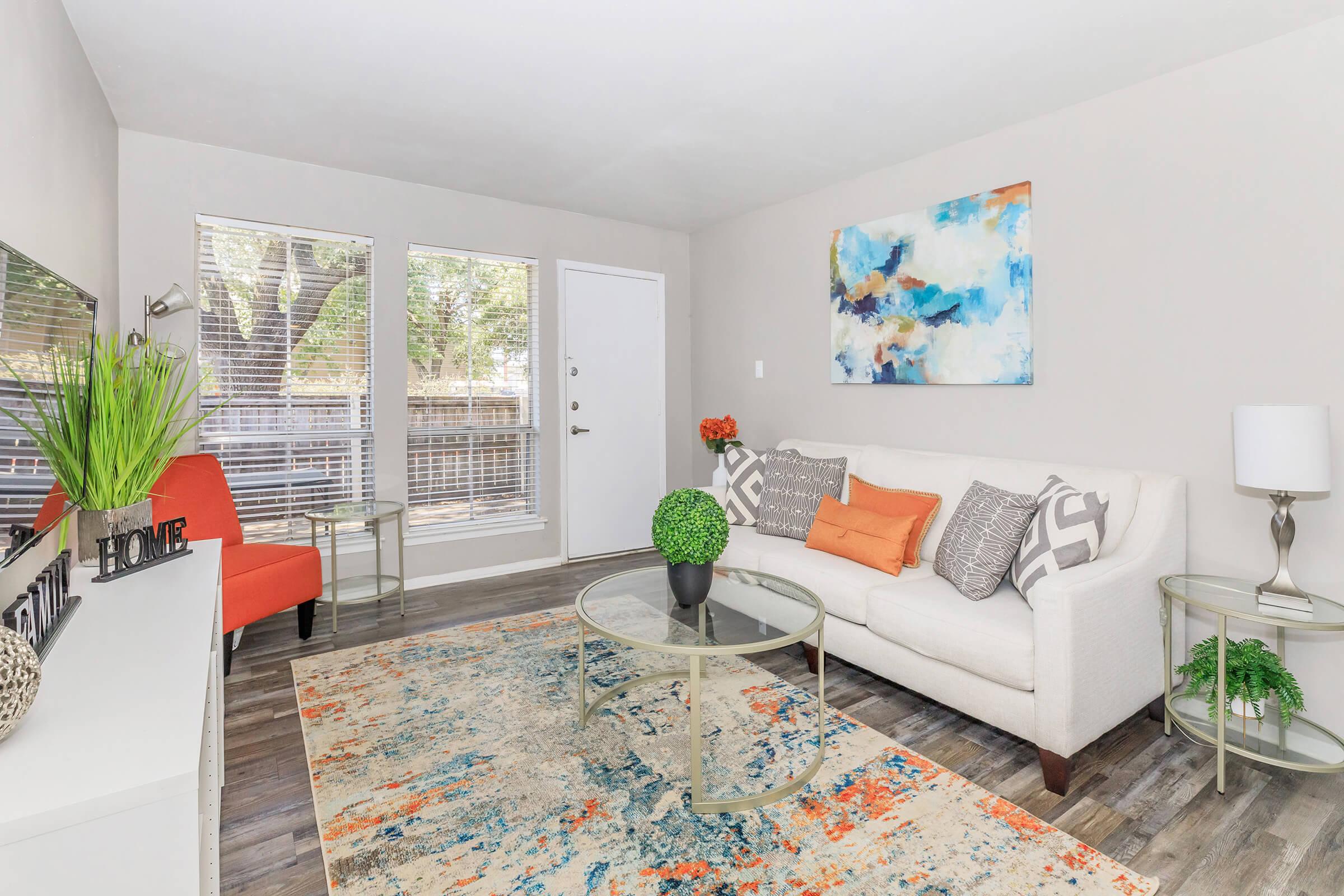 A cozy living room featuring a white couch with orange and gray decorative pillows, a round glass coffee table, and a colorful area rug. Large windows offer natural light, and there's a colorful abstract painting on the wall. An orange accent chair complements the space, along with green plants for added decor.