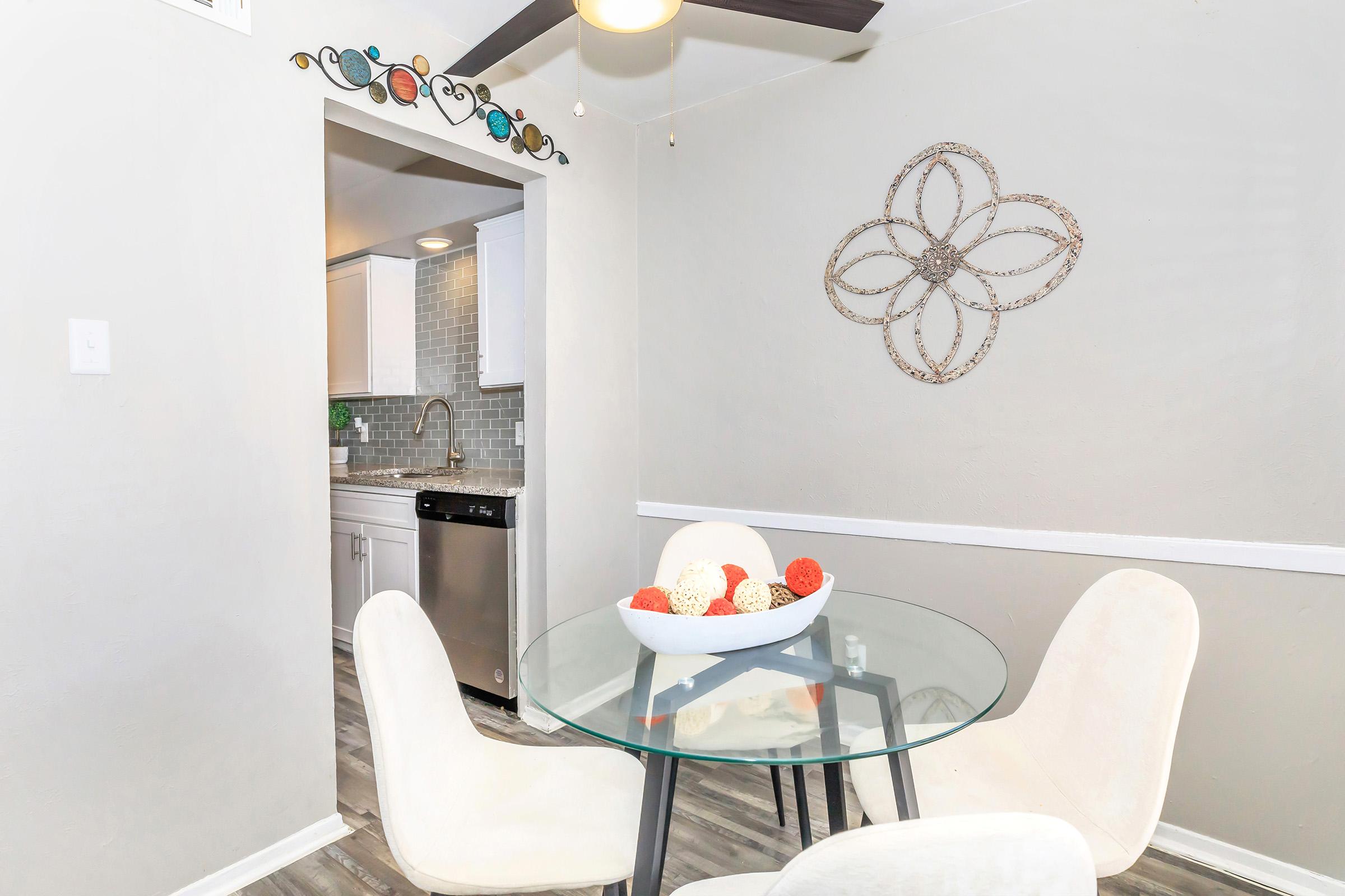A modern dining area featuring a round glass table with white chairs, a decorative bowl of colorful spheres, and a wall-mounted ornament. In the background, a kitchen with white cabinets and a stainless steel dishwasher is visible. The walls are painted in a neutral tone, enhancing the space's brightness.