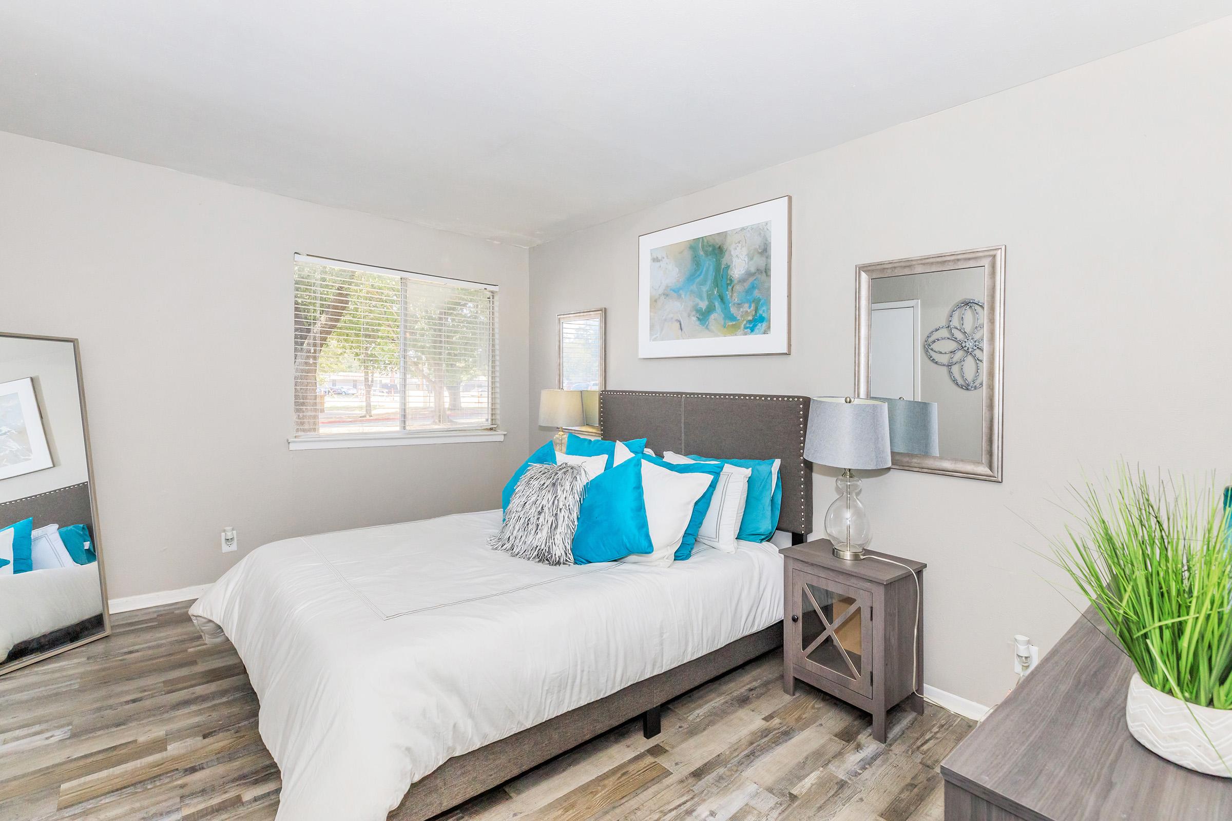 A cozy bedroom featuring a queen-sized bed with blue and white bedding, a decorative mirror, and a bedside table with a lamp. Natural light floods in from a window, and a green plant adds a touch of freshness to the decor. The wooden floor complements the modern aesthetic.