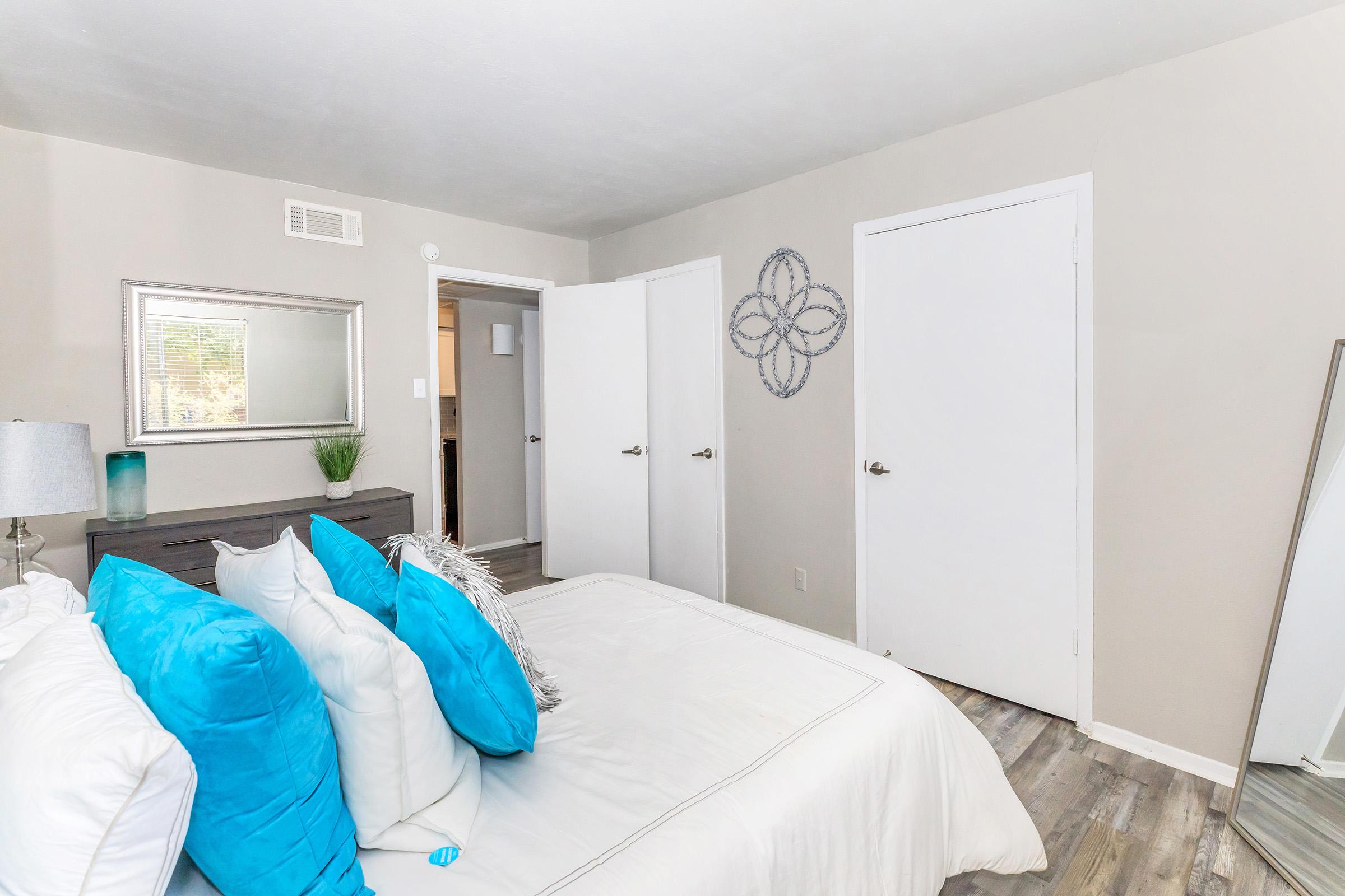 A modern bedroom featuring a neatly made bed with white linens and bright turquoise accent pillows. There's a stylish gray nightstand with a lamp, a large mirror, and a decorative wall art piece. Two doors lead to other rooms, and there's a potted plant in the corner, all set against light-colored walls and wooden flooring.