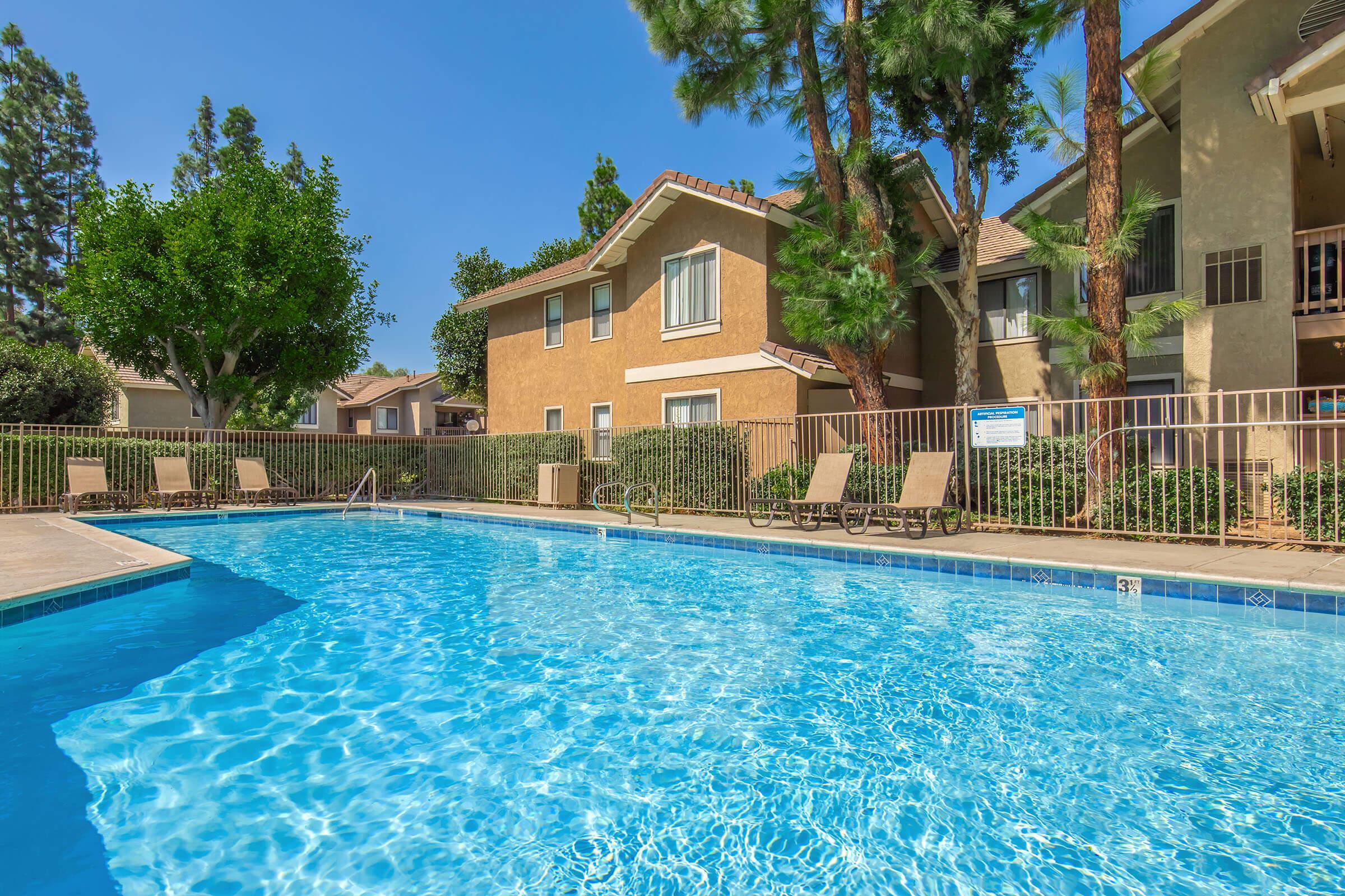 A sparkling blue swimming pool surrounded by lounge chairs, with two-story apartment buildings and tall trees in the background under a clear blue sky. The pool area is well-maintained, perfect for relaxation and leisure.