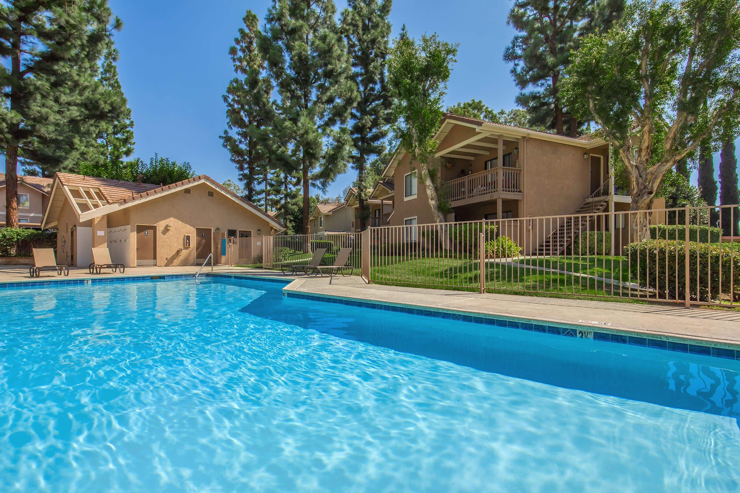 A clear blue swimming pool surrounded by lush green grass and tall trees. In the background, there is a beige building with balconies and a well-maintained landscaped area. Sunlight illuminates the scene, creating a bright and inviting atmosphere.