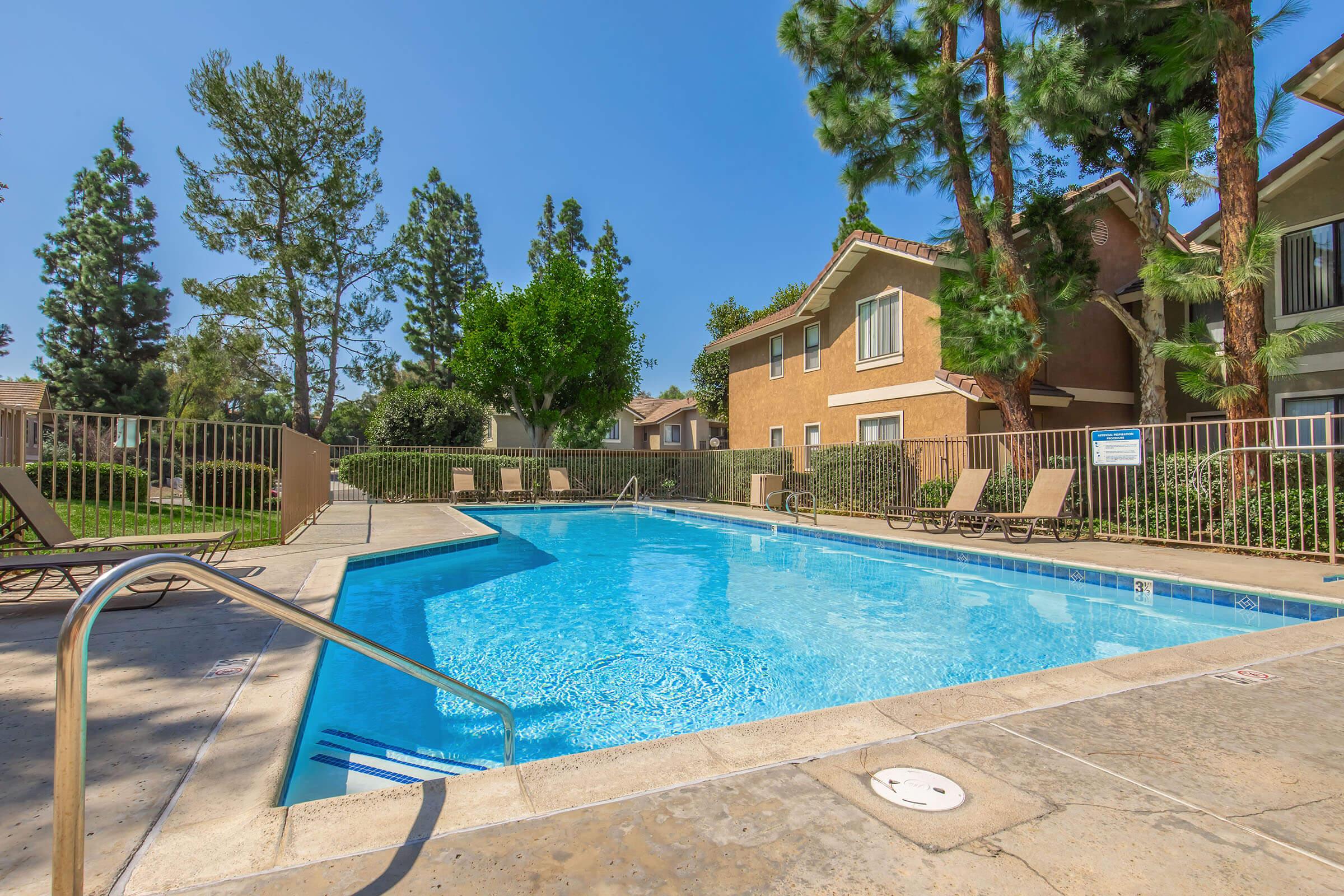Clear blue swimming pool surrounded by lounge chairs and lush greenery. Trees provide shade, and apartment buildings are visible in the background under a clear blue sky. The scene conveys a relaxing and inviting atmosphere typical of an outdoor pool area in a residential complex.