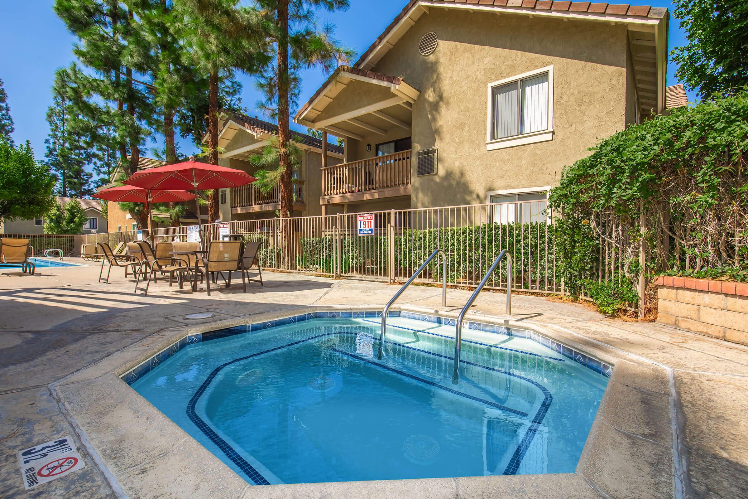 A clean, inviting hot tub beside a pool area, surrounded by lush greenery and tall trees. In the background, there are two residential buildings with balconies. A few lounge chairs and a large red umbrella provide shade, creating a relaxing outdoor environment.