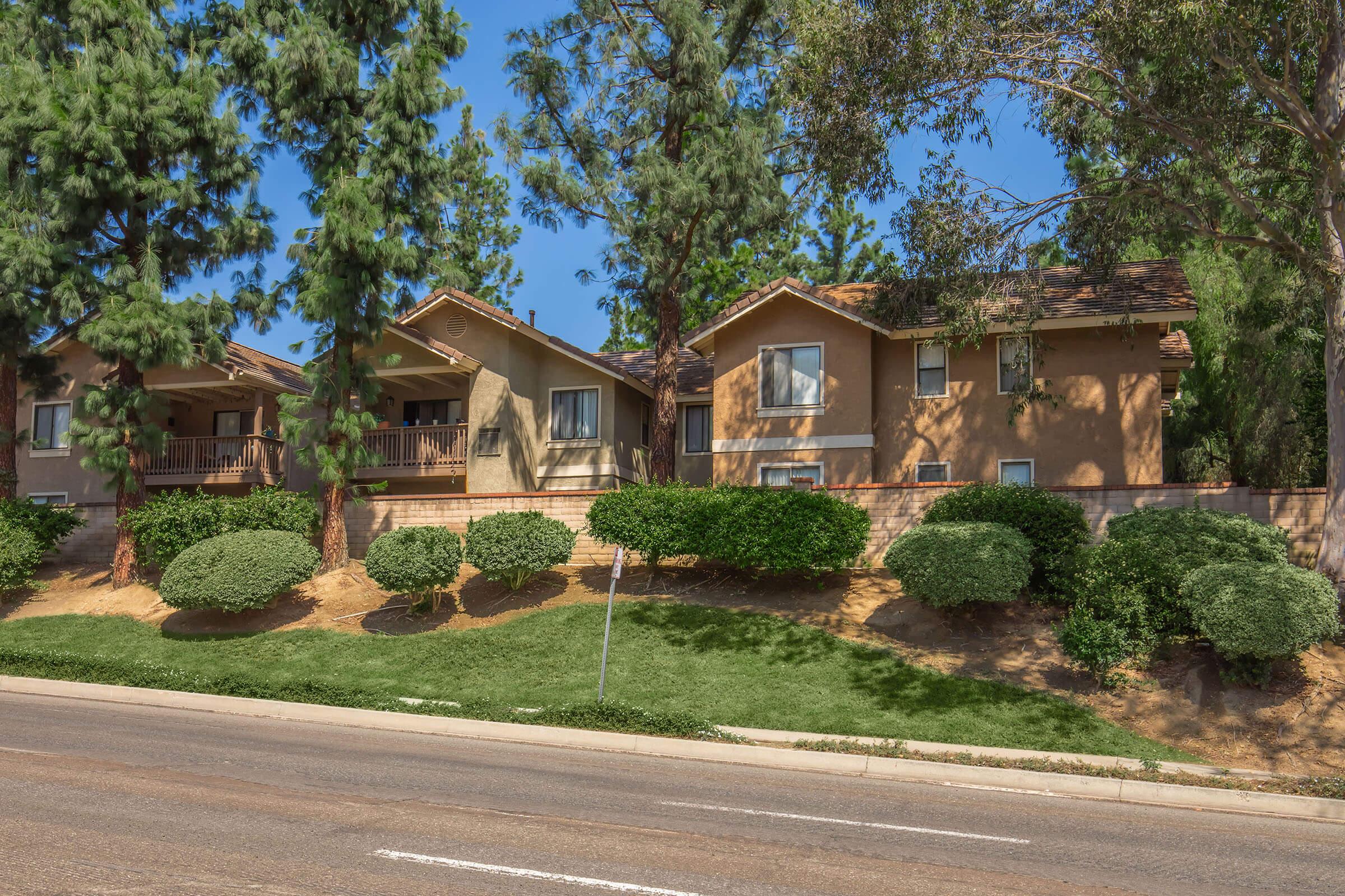 A row of residential houses situated on a hillside, surrounded by lush green trees. The homes have varied architectural styles and colors, with well-maintained landscaping in front. A quiet road runs alongside the houses, showcasing a peaceful suburban setting.