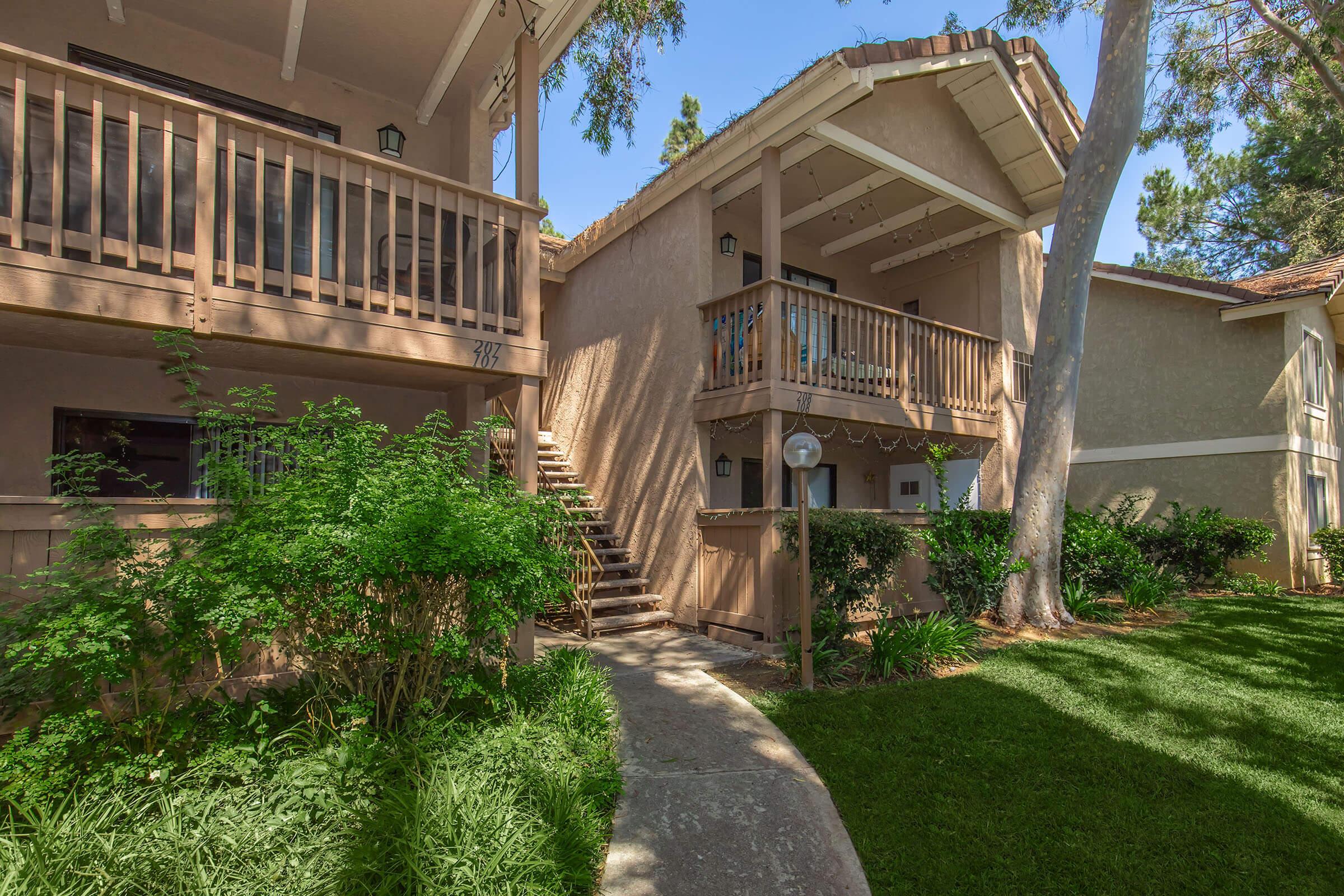 A well-maintained two-story building featuring a balcony, with a neatly landscaped pathway leading to the entrance. Lush green plants and trees adorn the surroundings, creating a peaceful atmosphere. The structure has a light-colored exterior with wooden accents and outdoor lighting fixtures.