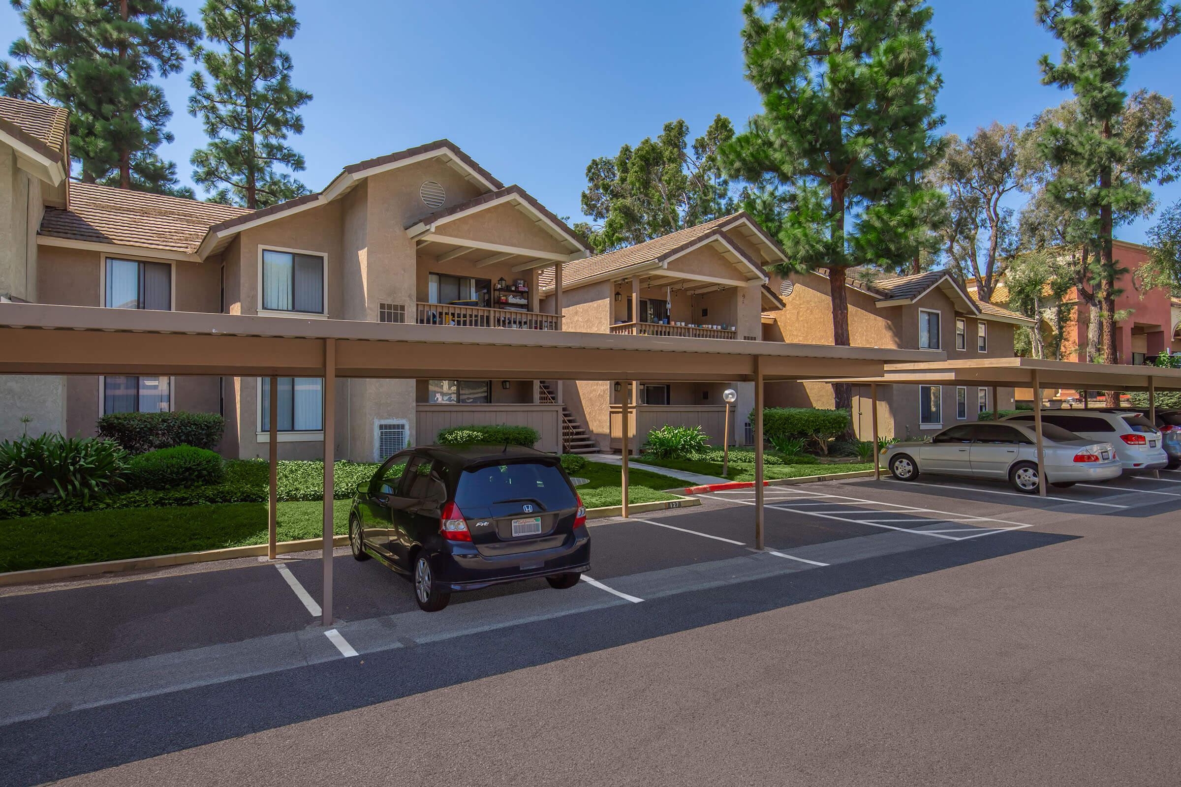 A view of a residential complex featuring several apartment buildings. There are covered parking spaces in the foreground, with a black car parked underneath a canopy. The area is surrounded by green trees and well-maintained landscaping, under a clear blue sky.
