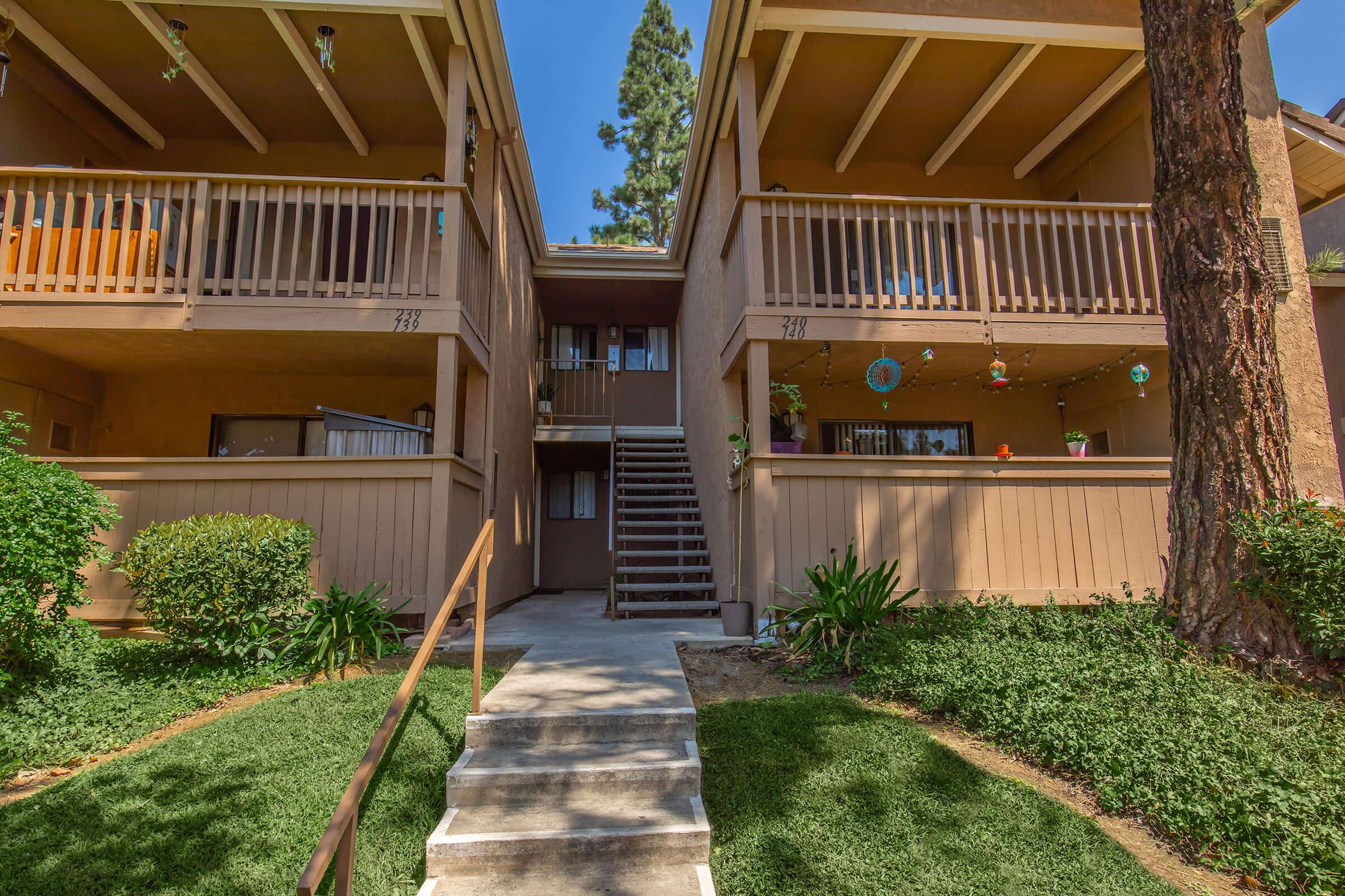 A view of a two-story apartment building with a front staircase leading to the upper level. The building features balconies with railings, surrounded by green grass and shrubs. Decorative items hang from the balconies, and tall trees are visible in the background under a clear blue sky.