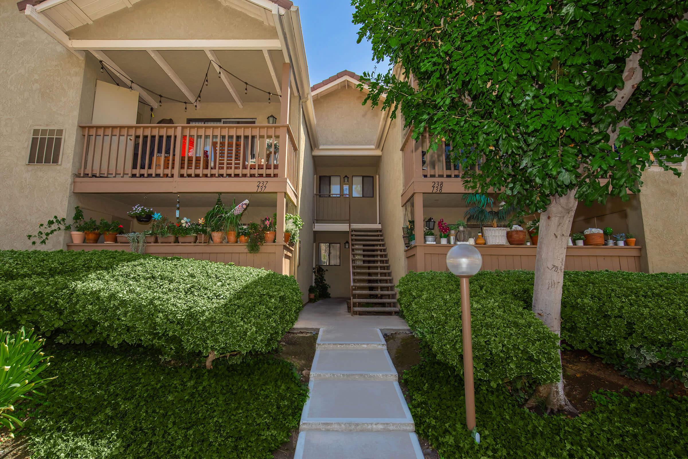 A well-maintained apartment building with a staircase leading to the upper floor. The ground floor features a walkway surrounded by neatly trimmed bushes, and several potted plants adorn the balconies. Bright blue sky visible overhead, adding a cheerful ambiance to the scene.