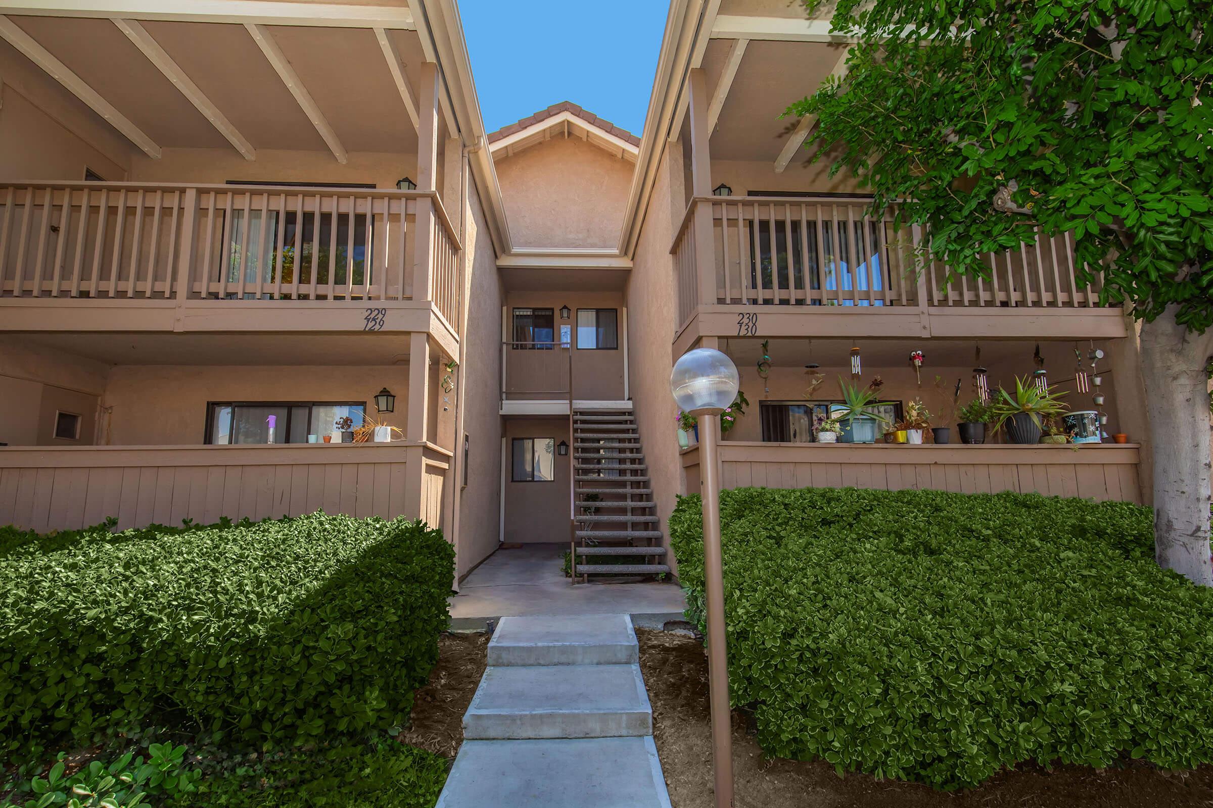 A view of a two-story apartment complex with a central staircase leading to upper balconies. The building features beige walls, wooden railings, and several potted plants on the balconies. Lush green shrubs are planted in front of the building, creating an inviting outdoor space under a clear blue sky.