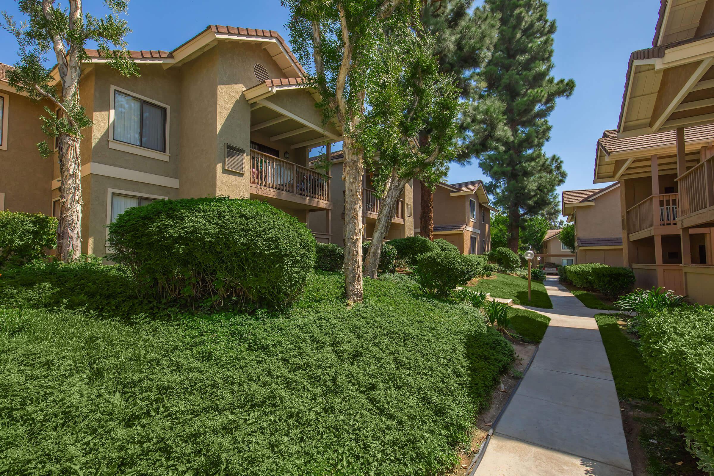 A peaceful residential area featuring two-story apartment buildings surrounded by well-maintained greenery and shrubs. A clean walkway meanders through the landscape, providing access to the buildings under a bright, clear blue sky. Sunlight highlights the lush environment.