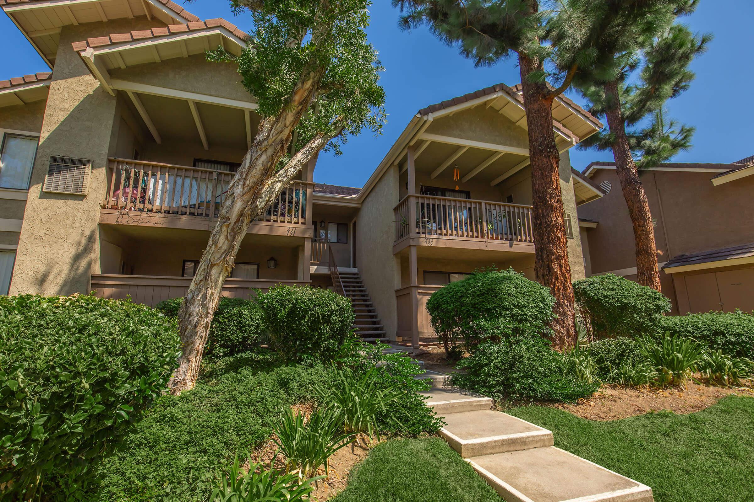 Two-story apartment buildings with wooden balconies surrounded by greenery, including shrubs and trees. A walkway leads to the entrance, and the sky is clear and blue, indicating a sunny day. The architectural style is modern, with a warm color palette.
