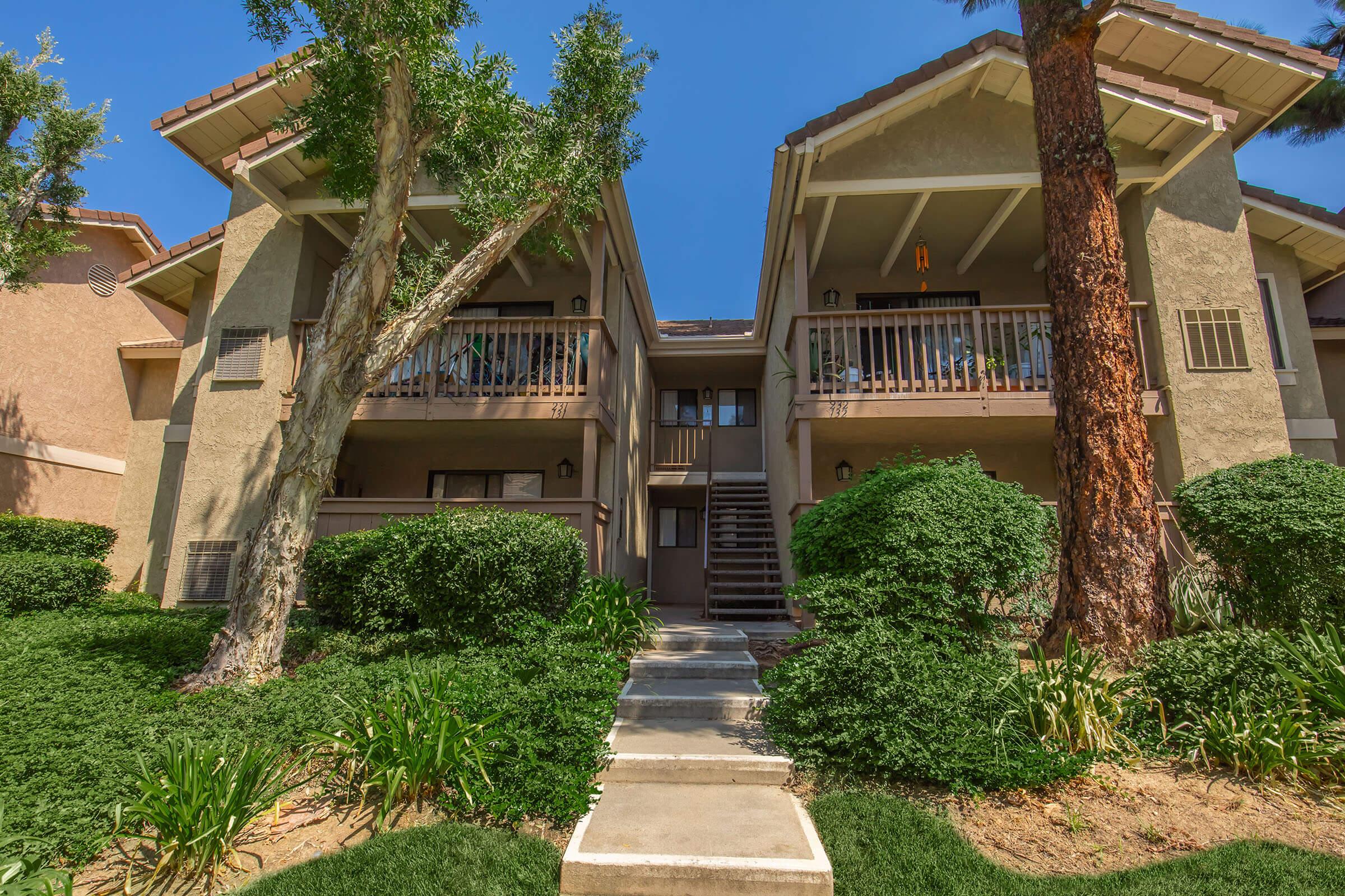 Two-story apartment building surrounded by lush greenery. The building features balconies on the upper level and steps leading to the entrance. Tall trees and well-maintained landscaping enhance the outdoor area, under a clear blue sky.