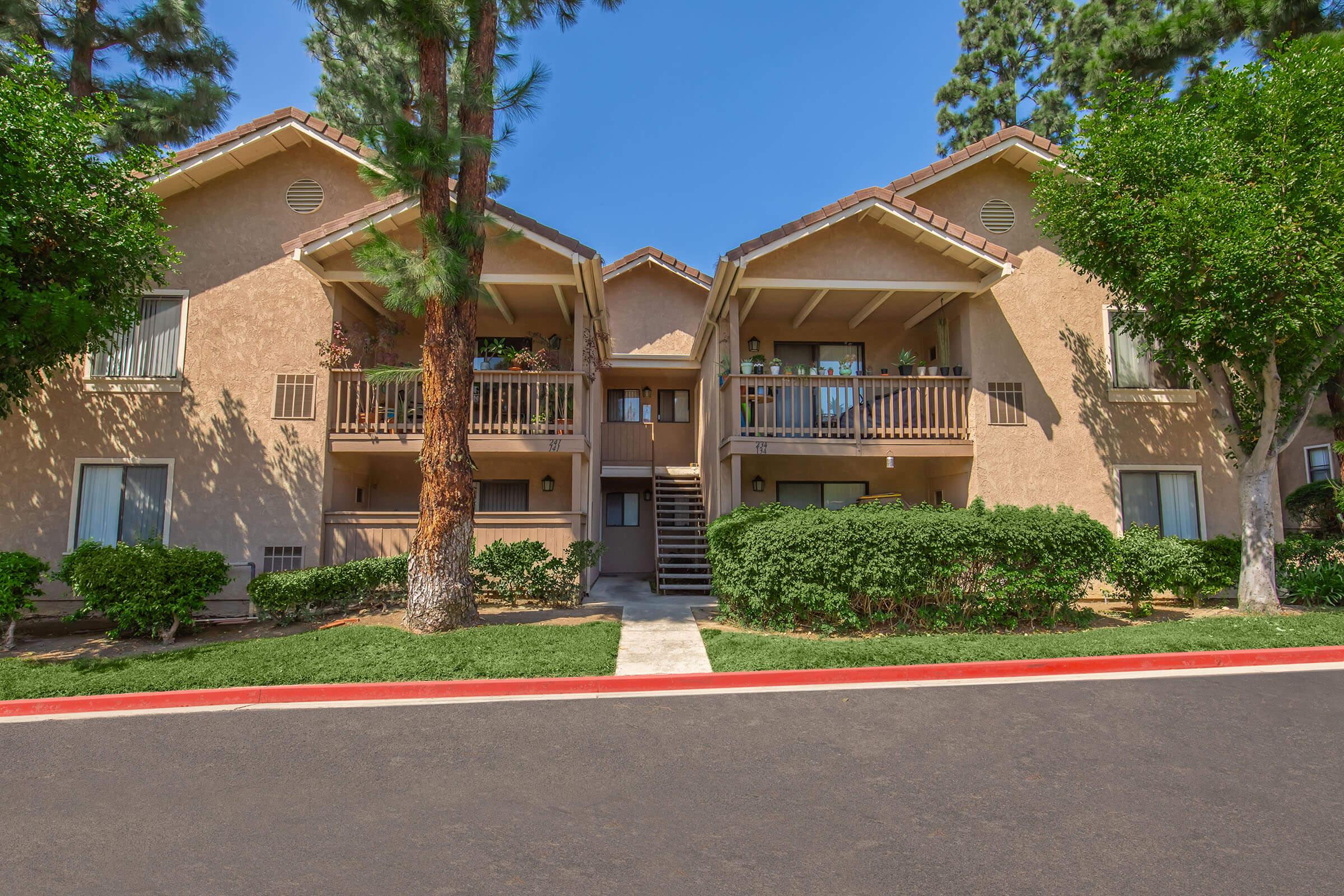 Two-story apartment building with a stucco exterior, featuring balconies on the second floor. The building is surrounded by neatly trimmed bushes and trees, with a clear blue sky overhead. A well-maintained pathway leads to the entrance, and a red line marks the edge of the adjacent parking area.