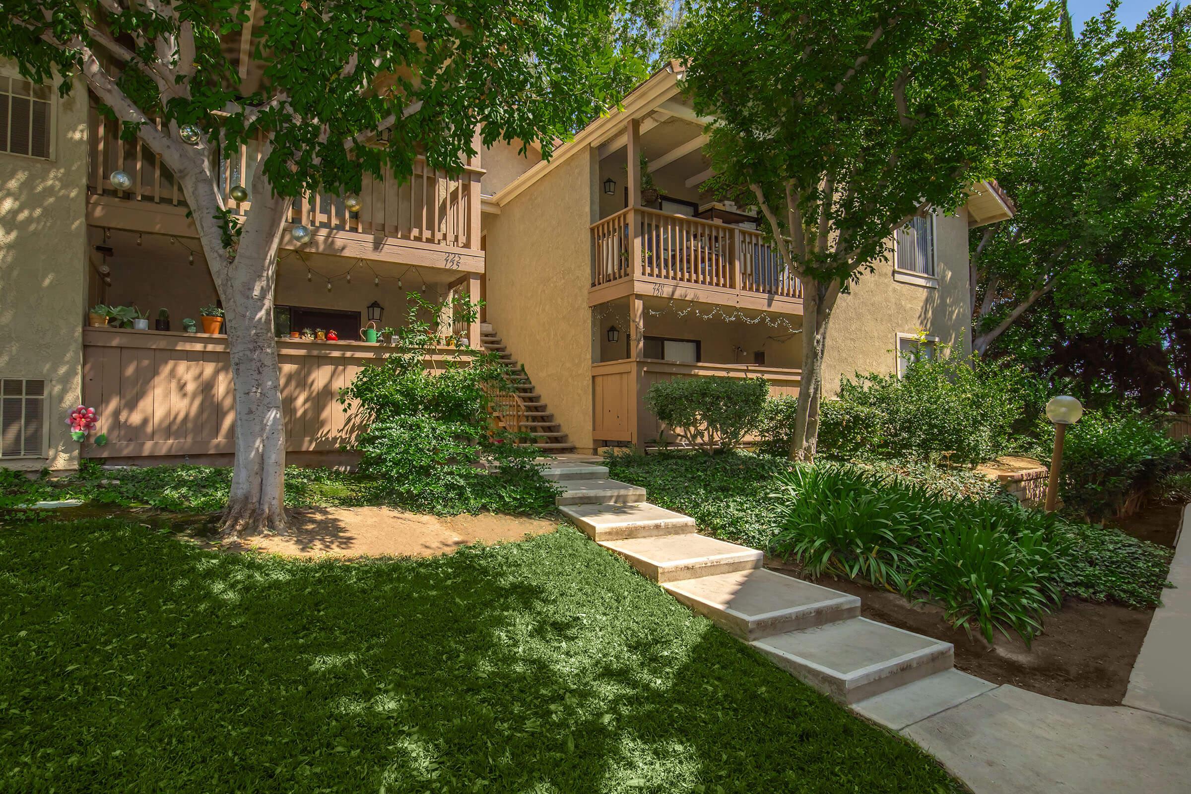 A landscaped garden featuring neatly trimmed grass, two trees, and a set of stone steps leading up to two multi-level residential buildings. The buildings have balconies adorned with plants, and the area is well-lit with a lamp post. Surrounding foliage adds to the greenery and charm of the setting.