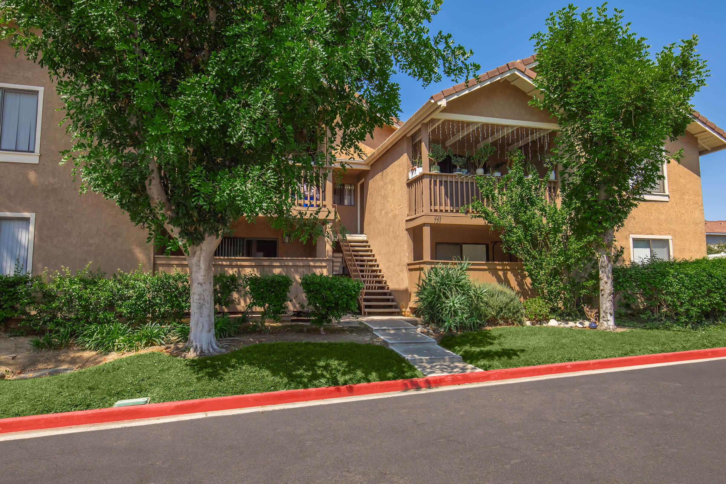 A two-story apartment building with a tan exterior and a balcony on the upper floor. Lush green trees and shrubs surround the building, and a stone pathway leads to the entrance. The scene features a blue sky and well-maintained landscaping, with a red-bordered pavement in the foreground.