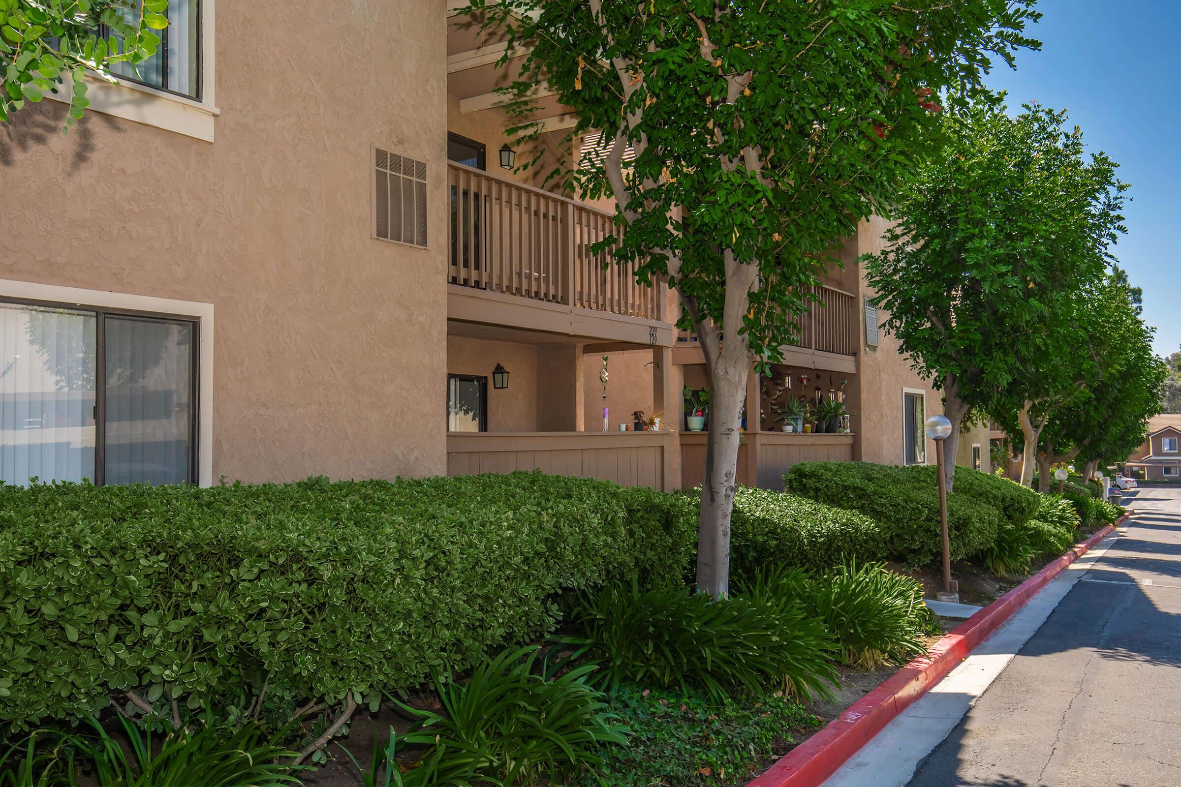 View of a residential building with a light beige exterior. The image shows two balconies adorned with potted plants. Lush green shrubs and small trees line the pathway, while a red curb runs along the side of the street, creating a well-maintained and inviting atmosphere. Bright blue sky above enhances the suburban setting.