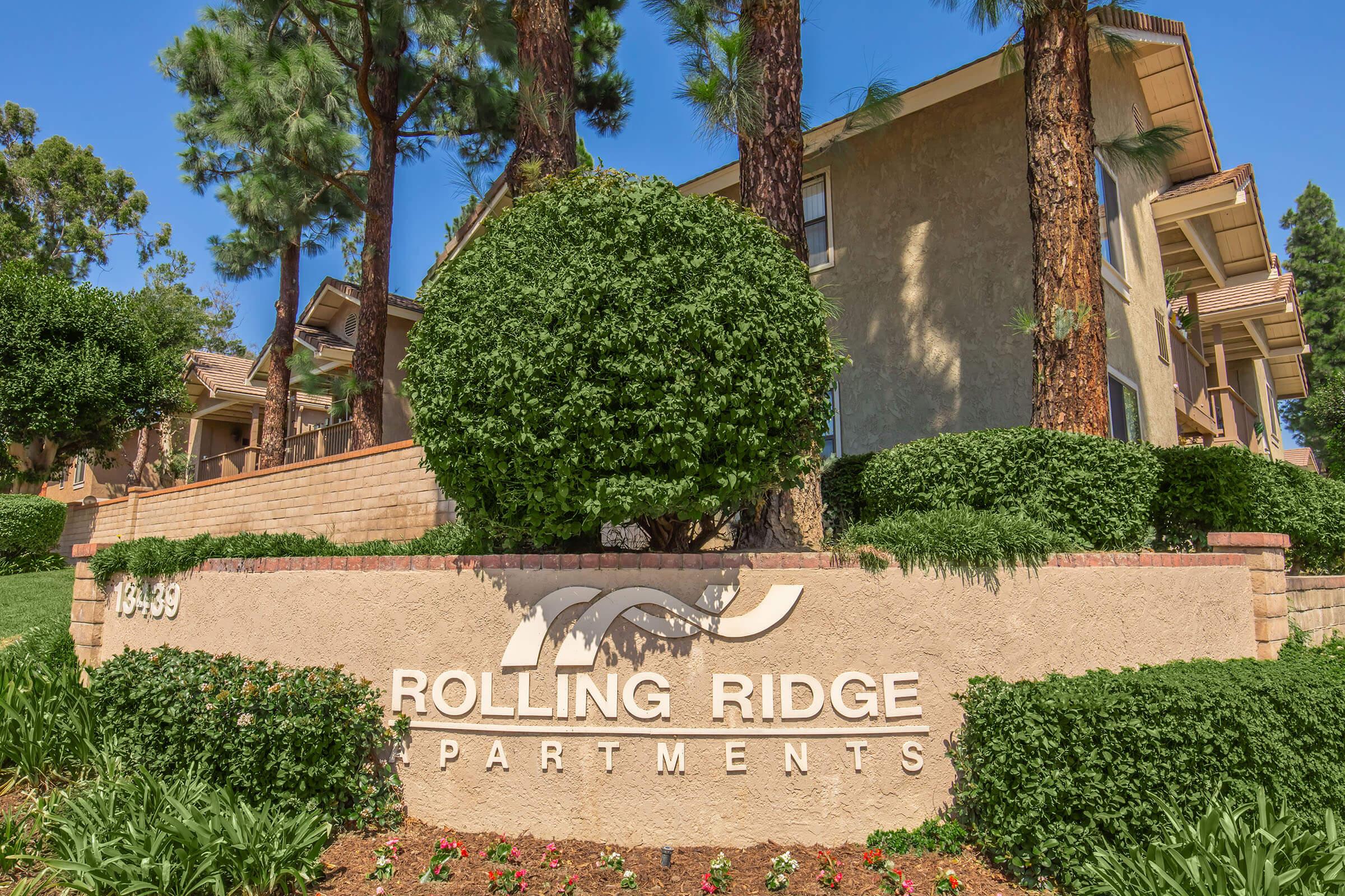 A well-maintained exterior view of the Rolling Ridge Apartments, featuring a sign with the text "ROLLING RIDGE APARTMENTS" in bold letters. The sign is surrounded by lush green landscaping, including well-trimmed bushes and flowering plants, with tall pine trees in the background under a clear blue sky.