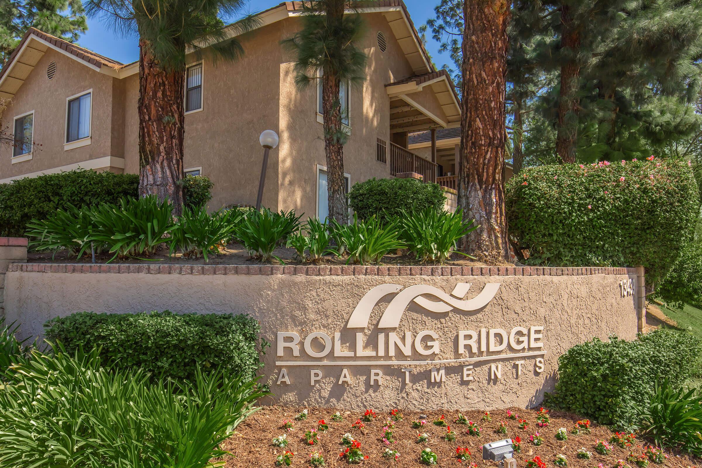 Sign for Rolling Ridge Apartments in front of a landscaped area with bushes and flowers. The apartment building is visible in the background, surrounded by tall pine trees. Clear blue sky overhead enhances the sunny atmosphere.