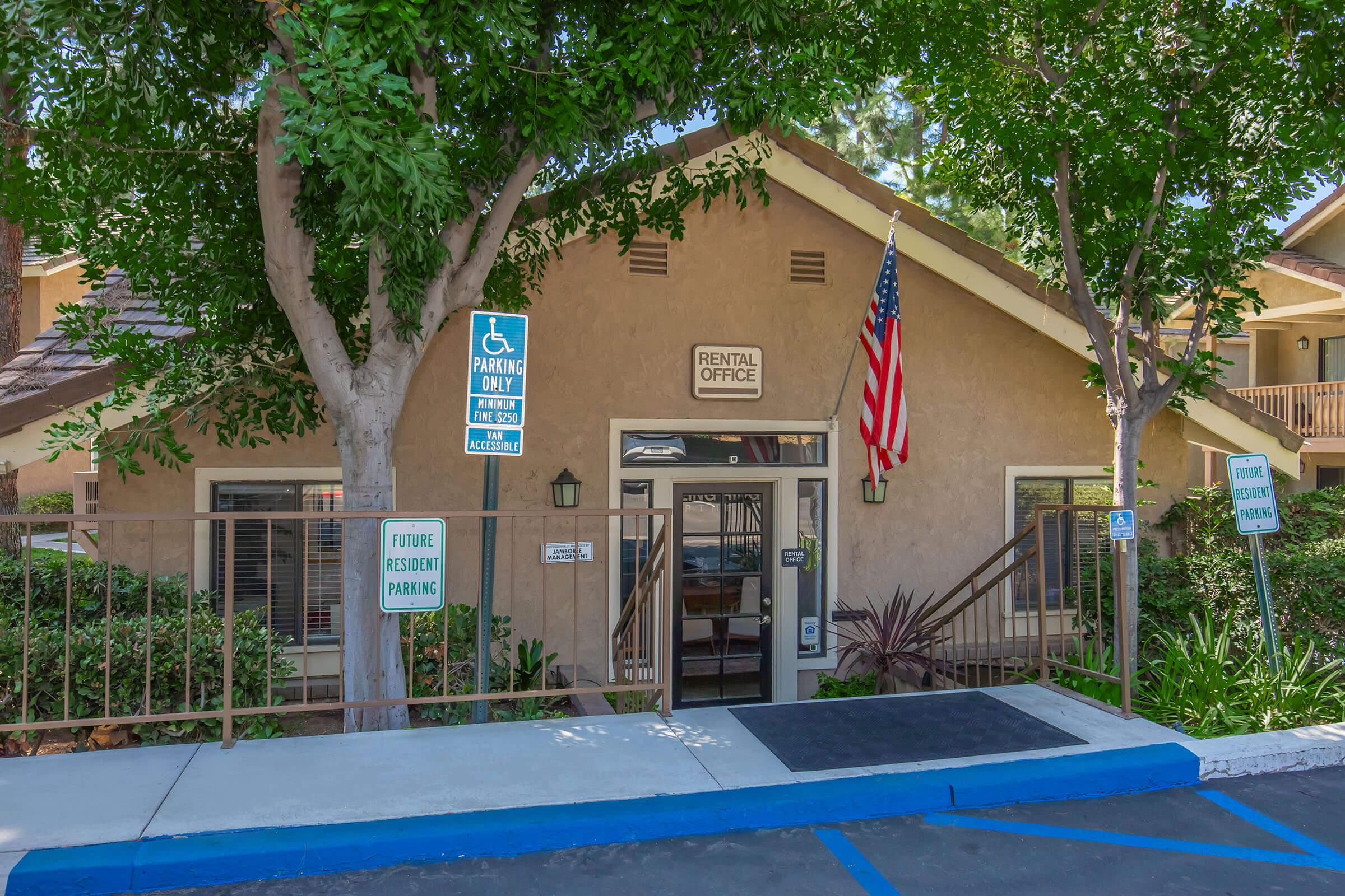 Exterior view of a rental office building surrounded by greenery, featuring large windows and a set of stairs leading to the entrance. A flag is displayed on the front, and parking signs indicate designated areas. The building has a welcoming appearance with a pathway leading up to the door.