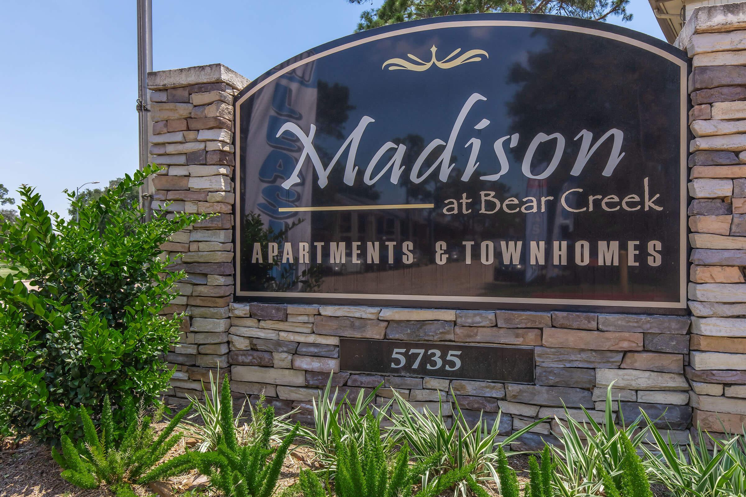Sign for "Madison at Bear Creek," featuring the name in elegant script above "Apartments & Townhomes." The sign is framed by a stone structure, with green landscaping in front, located at address 5735.