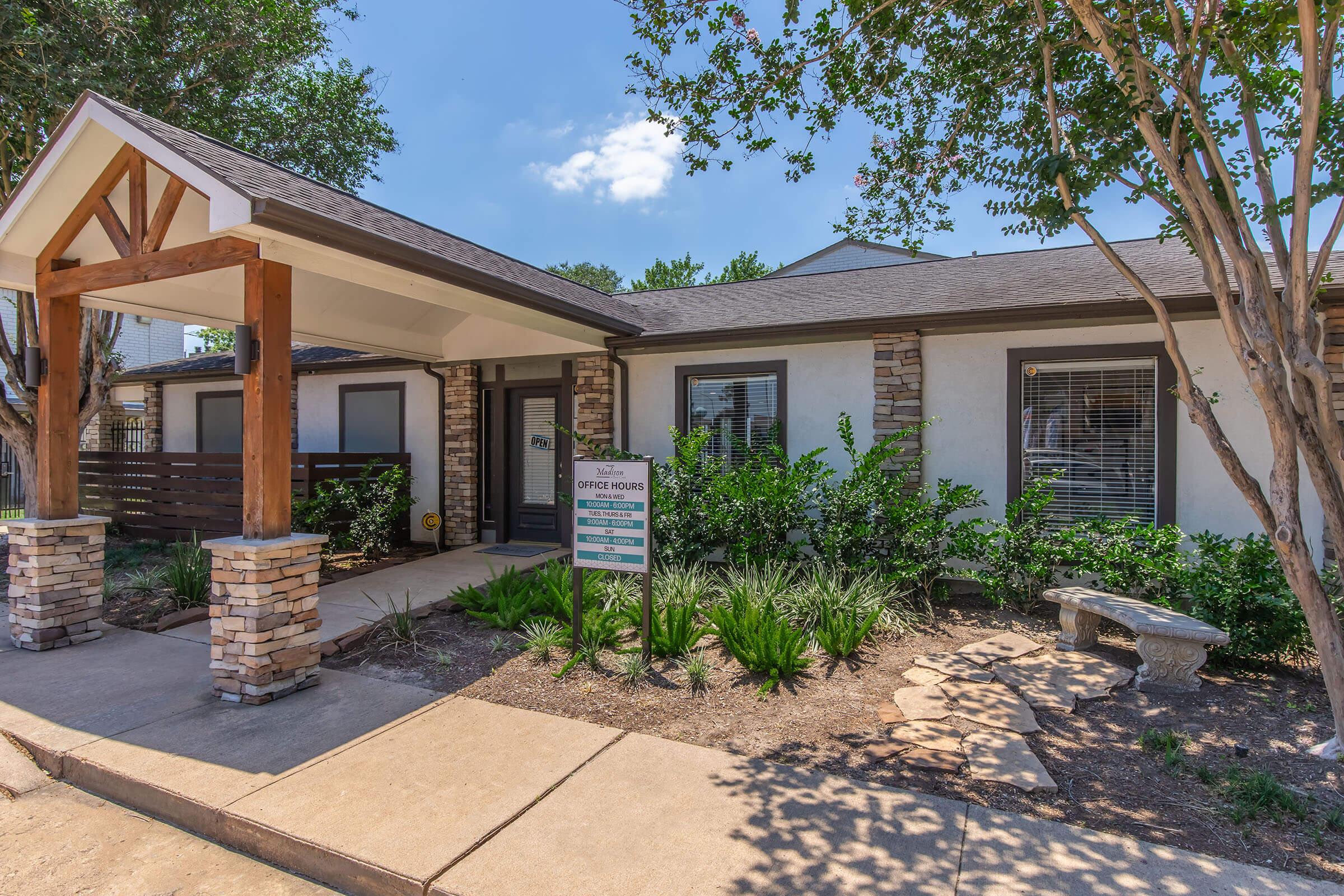 A modern office building with a welcoming entrance featuring wooden beams and stone pillars. The front yard is landscaped with greenery and a bench. A sign indicating "Office Hours" is displayed prominently. The sky is clear, and the scene is bright and inviting.