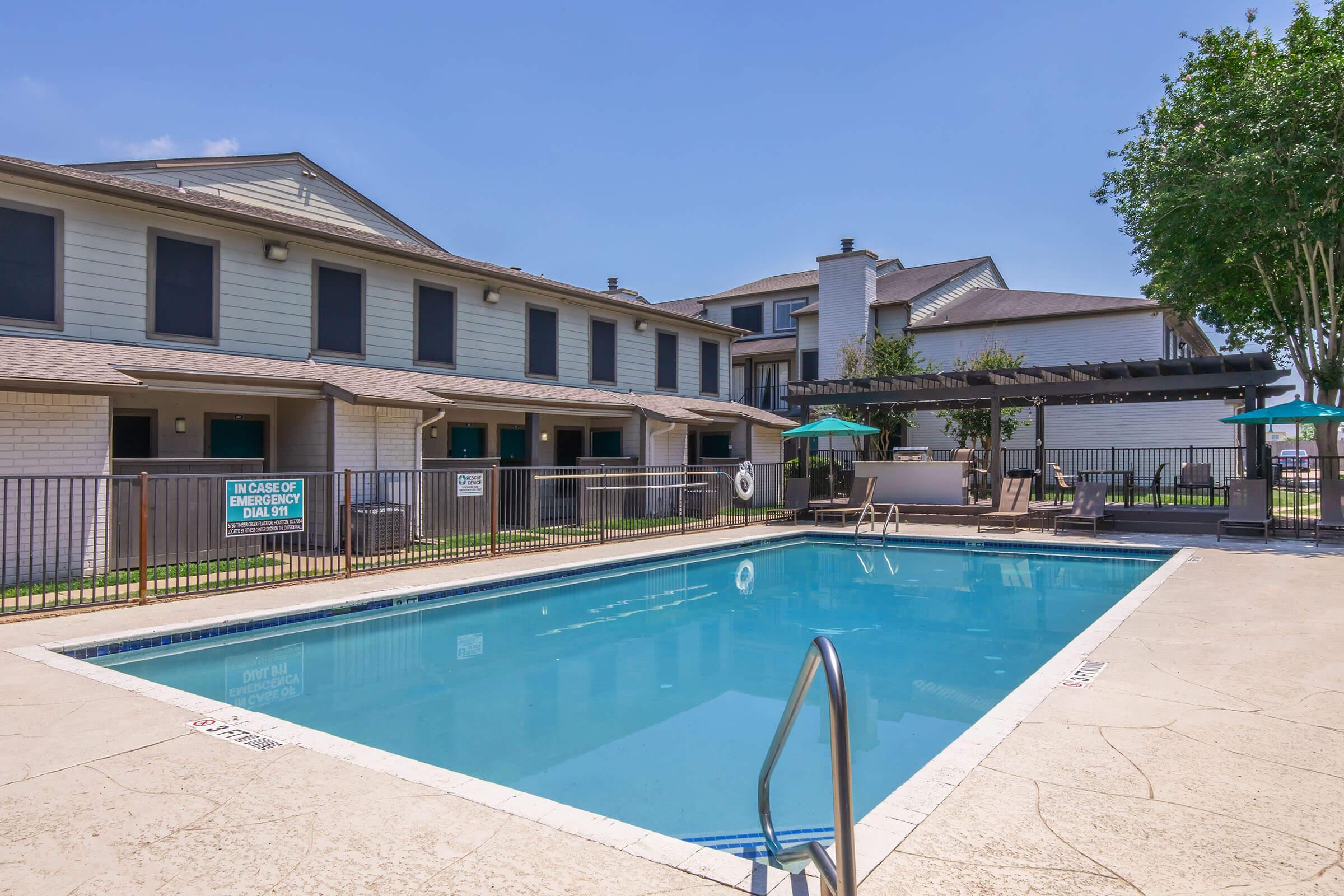 A clear swimming pool surrounded by a paved area, with lounge chairs and umbrellas. In the background, there are two multi-story residential buildings with balconies. The sky is blue and sunny, and there are trees nearby, providing a pleasant outdoor atmosphere.