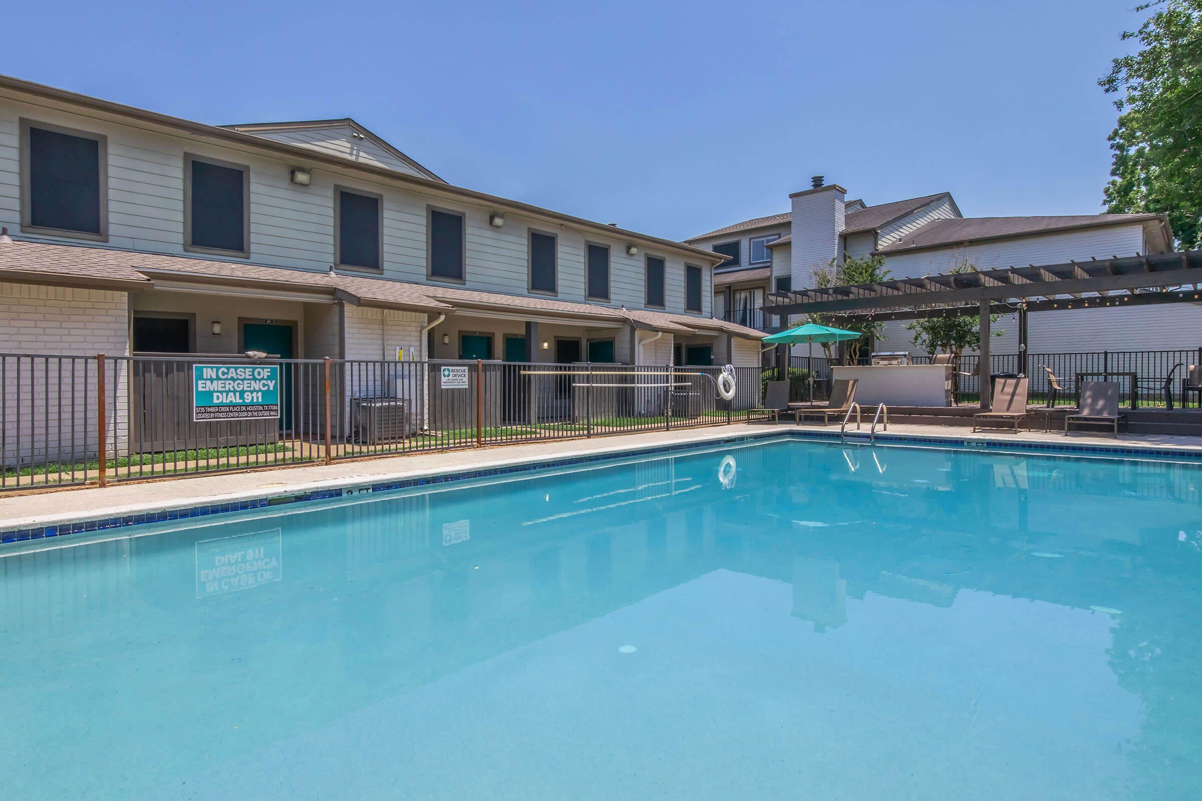 A swimming pool surrounded by a fenced area, with lounge chairs and an umbrella on one side. In the background, there are two multi-story residential buildings with windows and a pergola. A sign near the pool indicates emergency contact information. The sky is clear, and it's a sunny day.