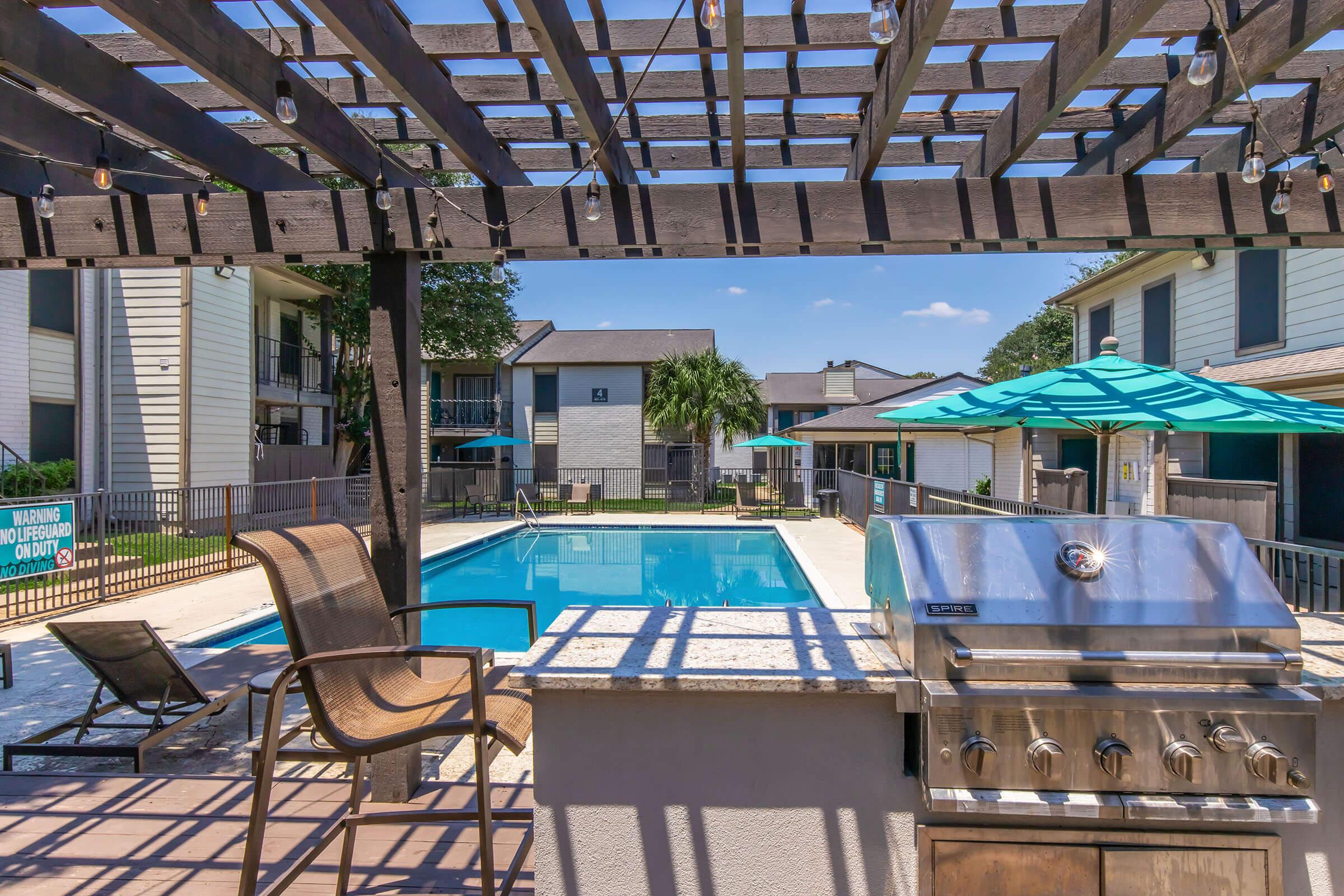 A serene outdoor pool area featuring a crystal-clear swimming pool surrounded by lounge chairs. A shaded patio with a wooden pergola and a grill offers a space for relaxation and dining. Umbrellas provide shade, and nearby buildings create a cozy community atmosphere under a clear blue sky.