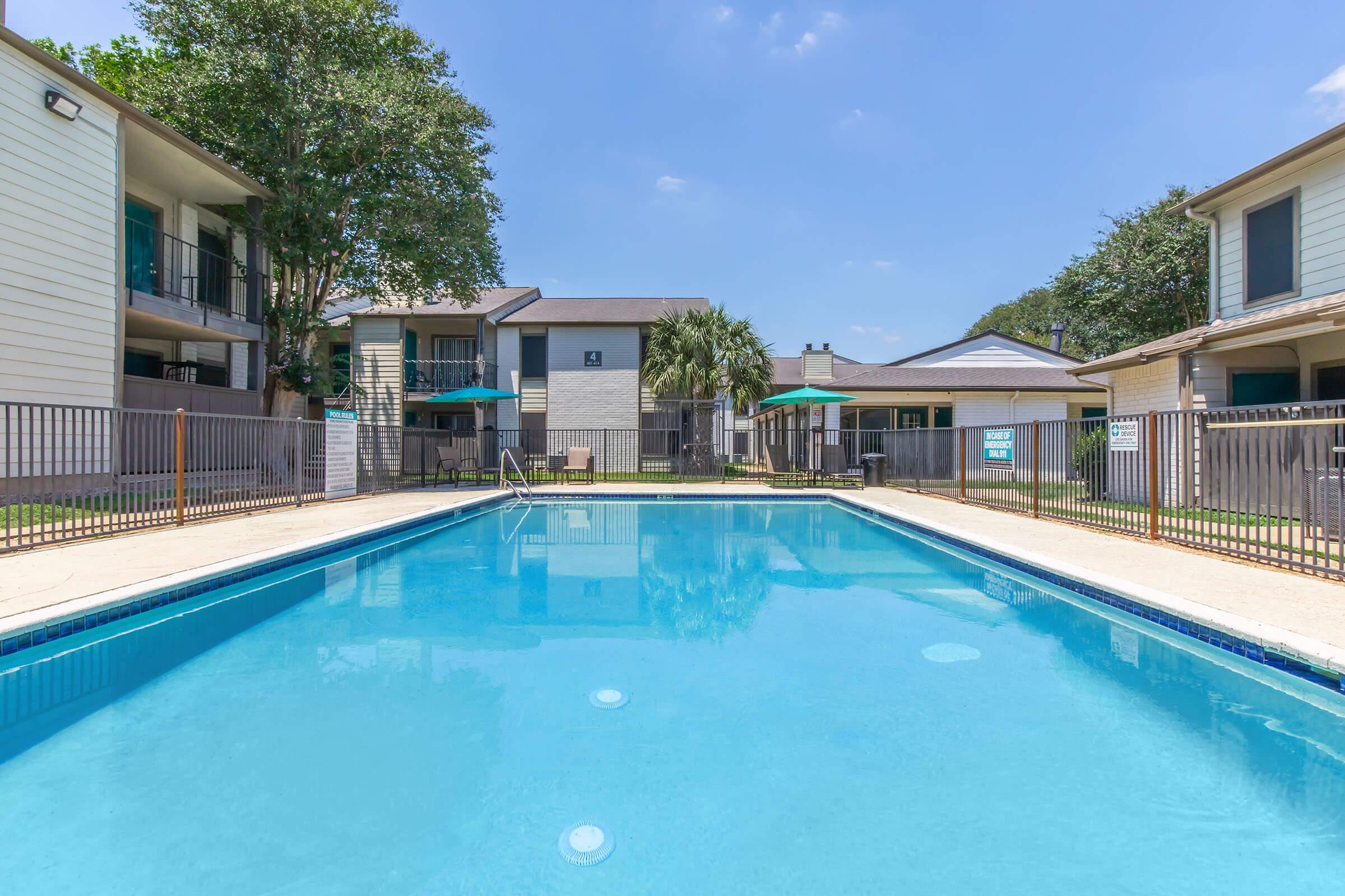 A clear swimming pool surrounded by a fenced area, with lounge chairs and umbrellas on the side. Apartment buildings with balconies are visible in the background, set against a blue sky with a few clouds. The scene is bright and inviting, suggesting a relaxing outdoor atmosphere.