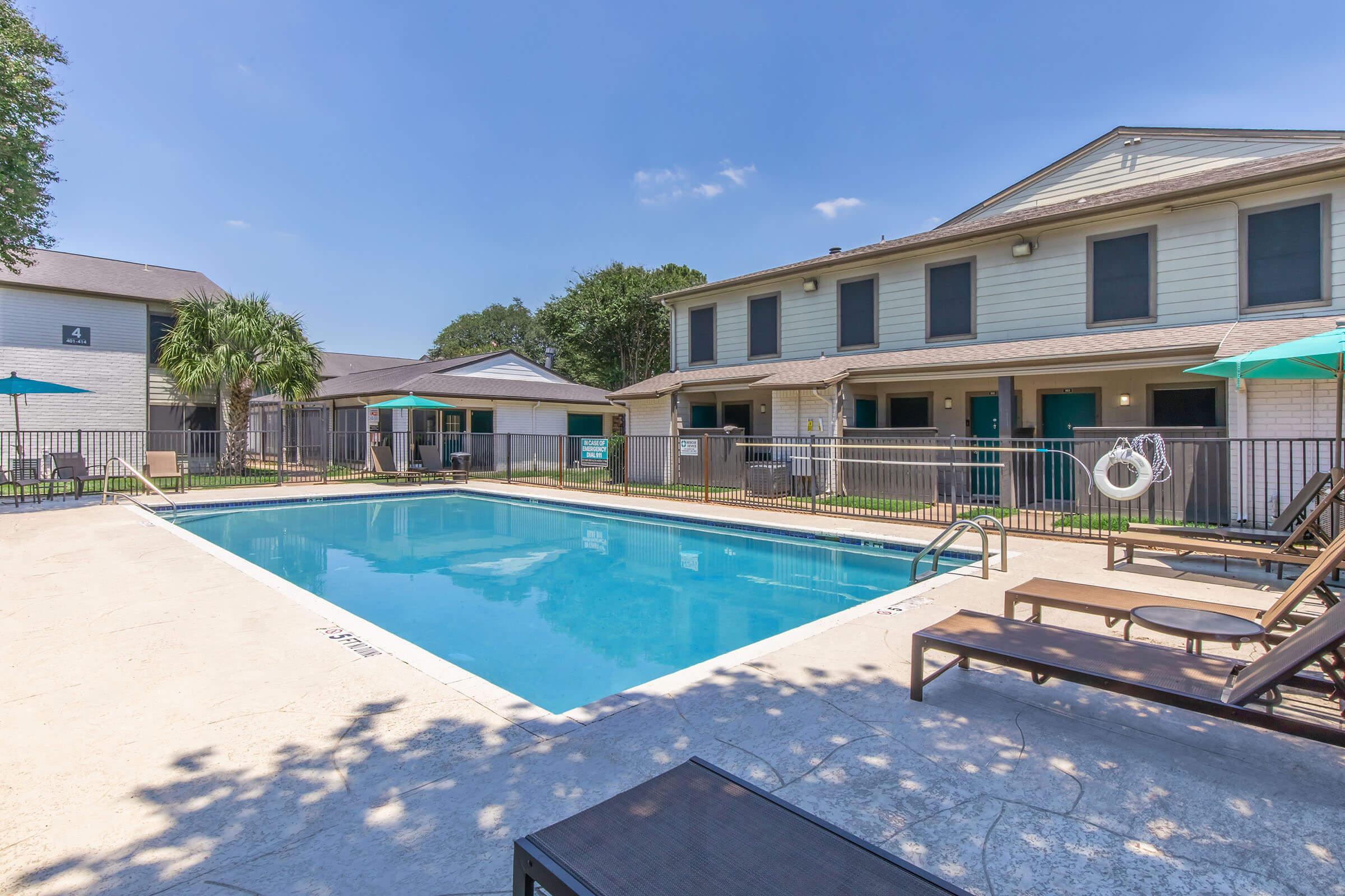 A residential area featuring a clear swimming pool surrounded by lawn chairs, umbrellas, and a fence. Nearby, there are two-story buildings with multiple units, lush greenery, and a sunny sky overhead.