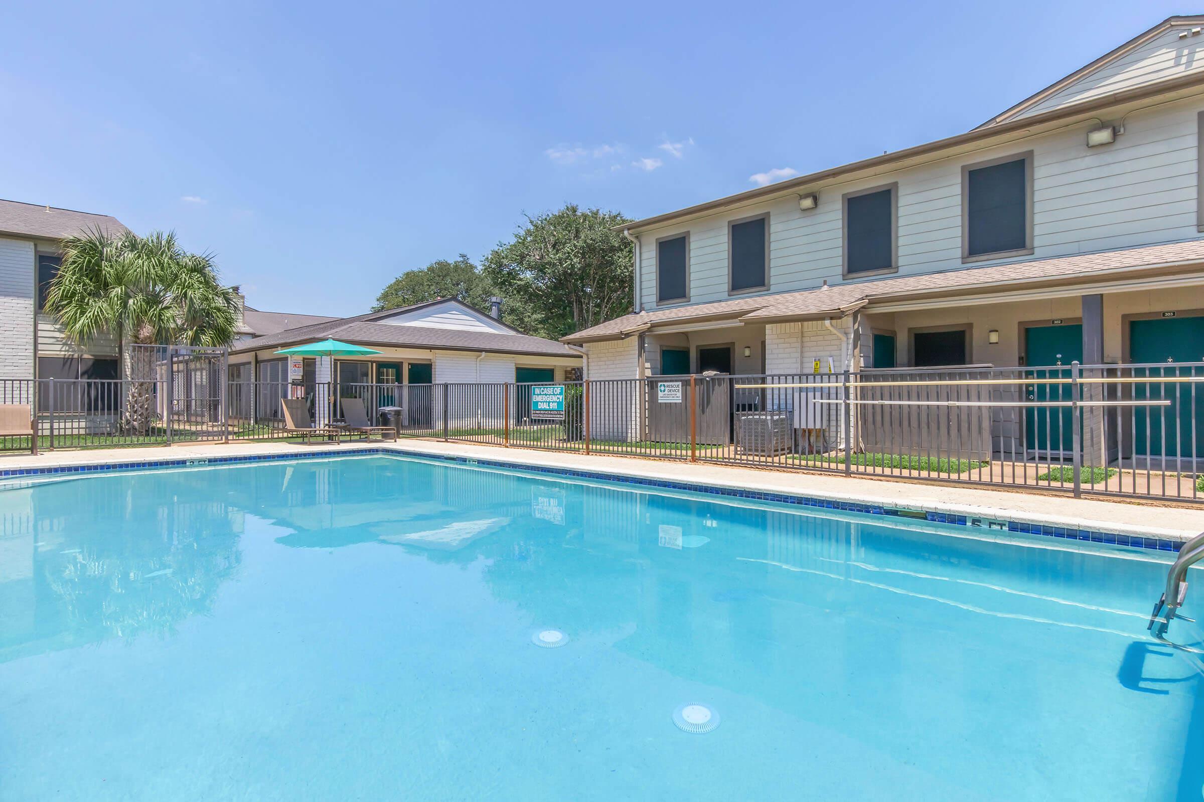 A clear blue swimming pool in the foreground with a poolside fence. In the background, two multi-story residential buildings are visible, along with a shaded seating area. The sky is bright and clear, indicating nice weather. Green trees can be seen in the vicinity.