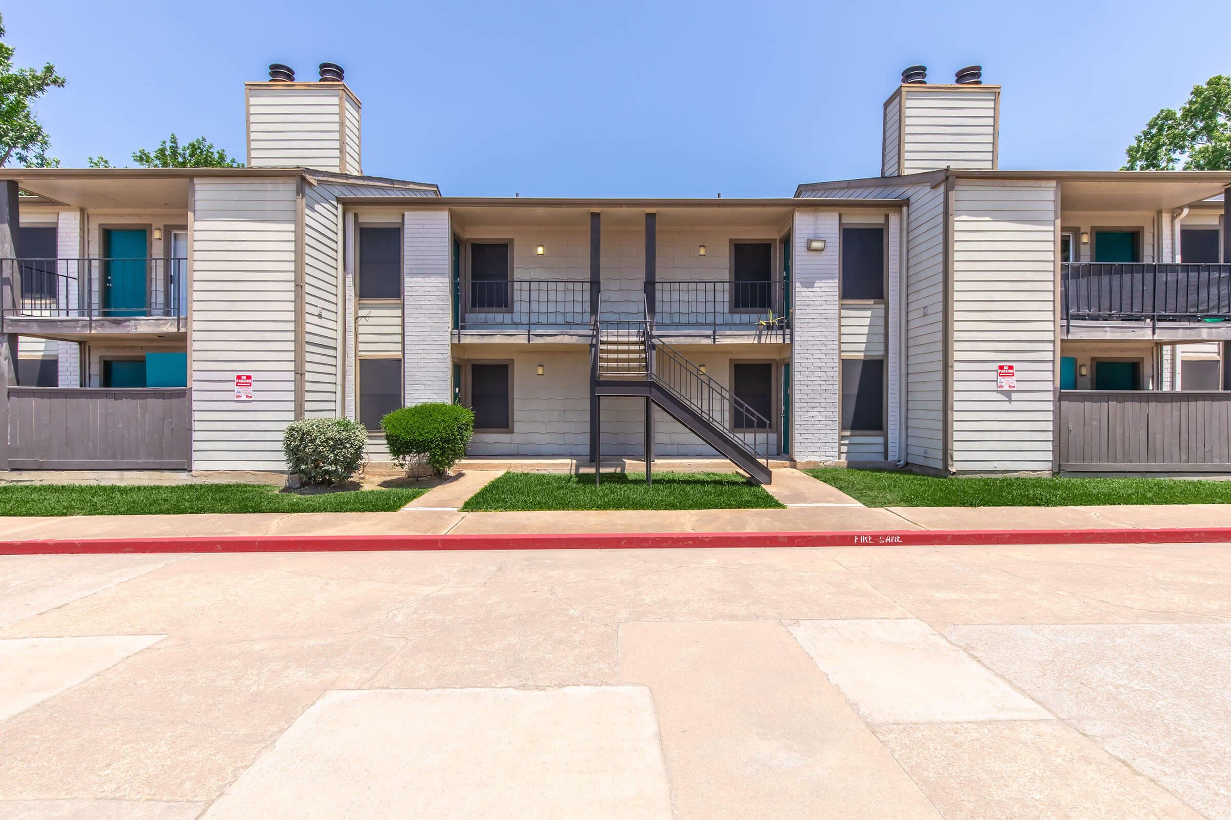 A two-story apartment building with a gray exterior and two staircases leading to the second floor. There are balconies on the upper level, a small grassy area in front, and a red curb by the entrance. The sky is clear and blue, providing a bright and inviting atmosphere.
