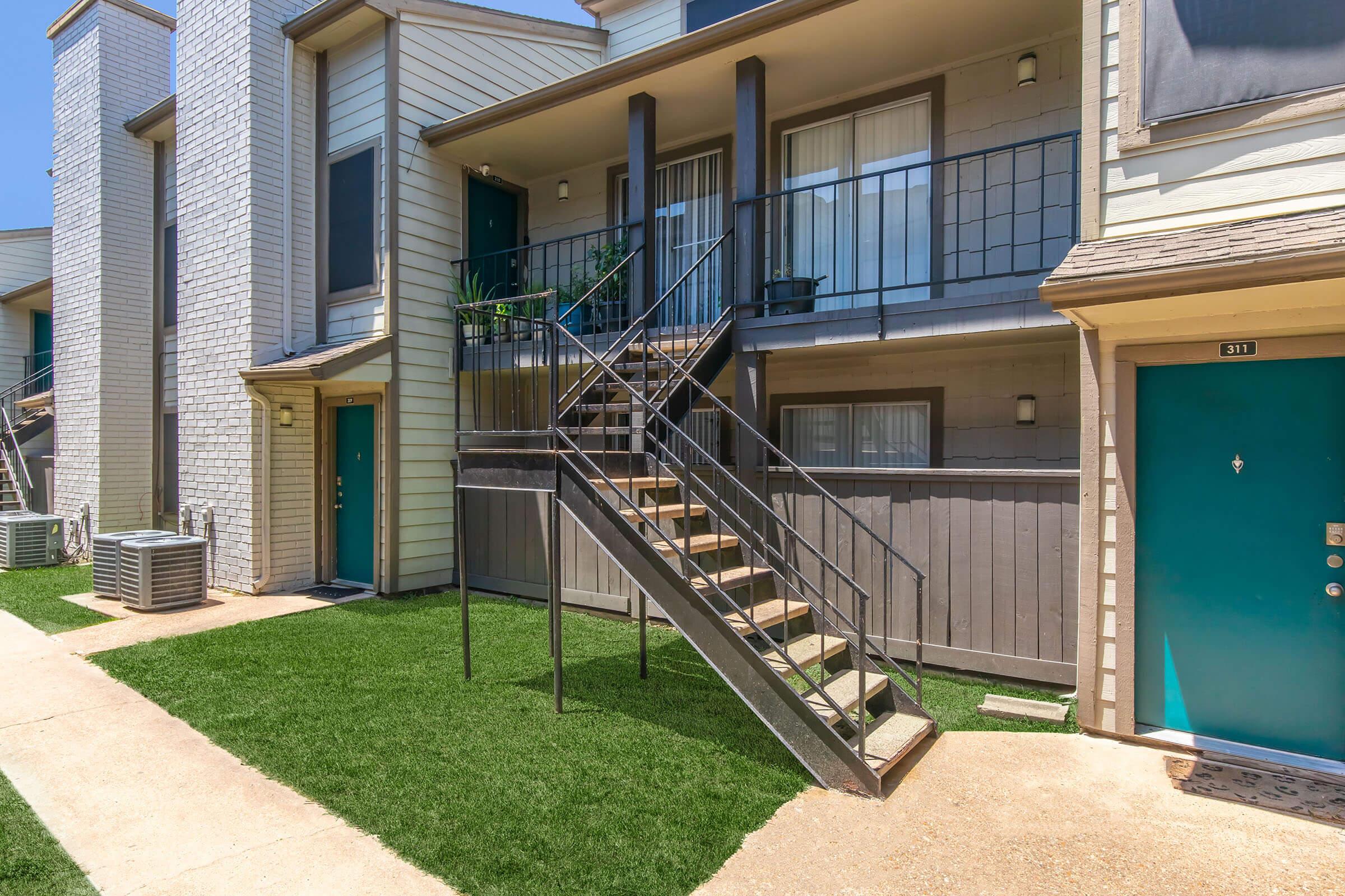 Exterior view of a multi-story apartment building with a staircase leading to a second-floor balcony. The building features a combination of light and dark siding, green doors, and a well-maintained lawn area. Air conditioning units are visible on the ground level.