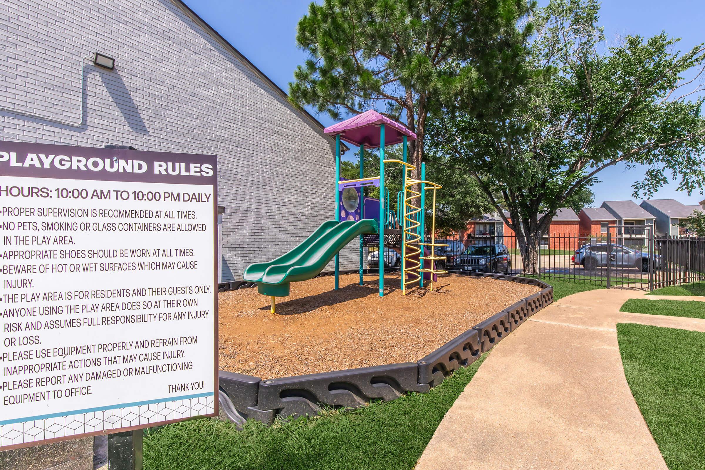 Playground area featuring a multi-colored play structure with slides, surrounded by safety surfacing. A sign lists rules, stating proper supervision is required, no pets or glass containers are allowed, and visitors must be aware of surfaces to prevent injuries. The area is well-maintained and enclosed by a fence.