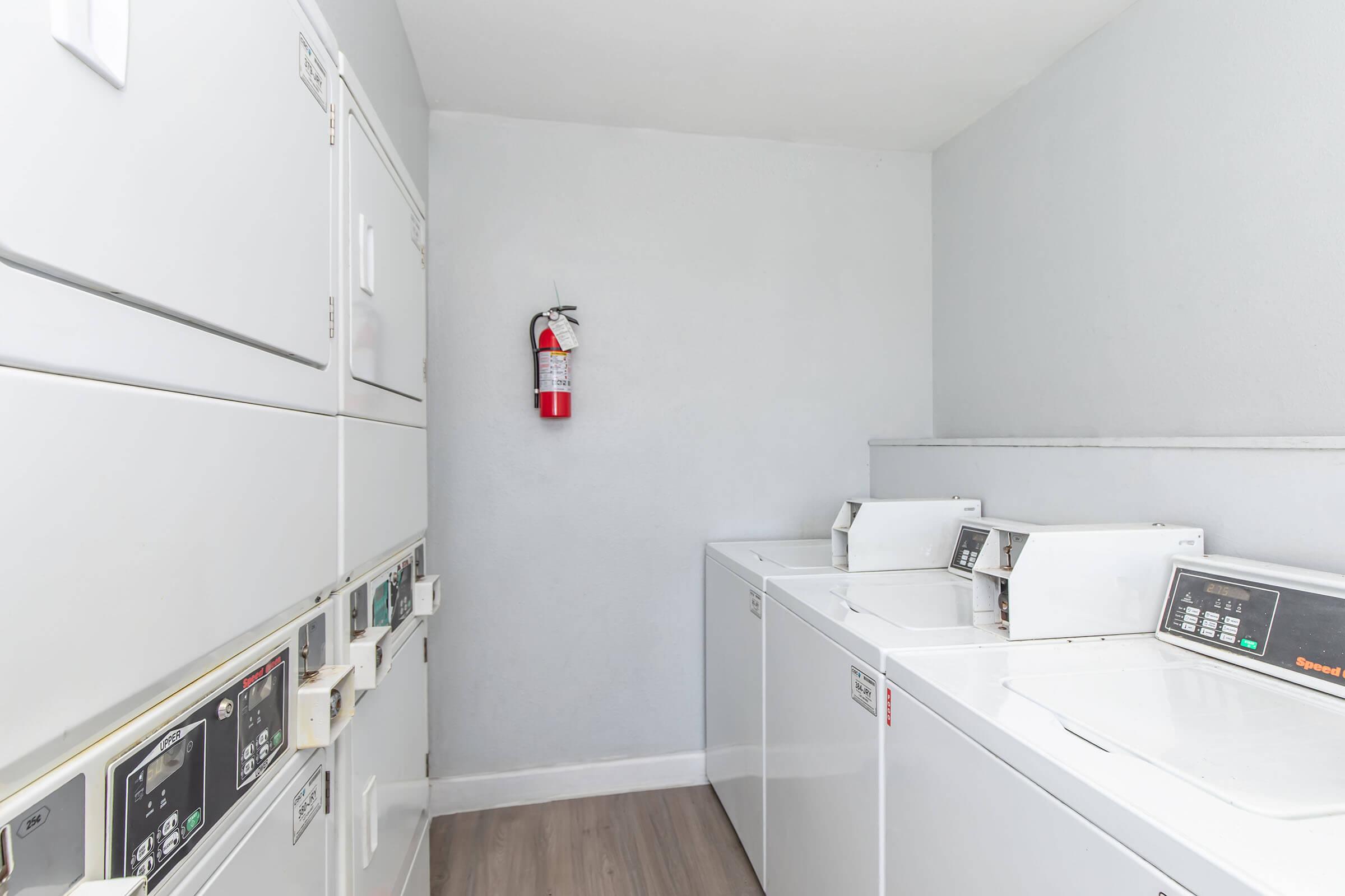A laundry room with several stacked washing machines and dryers. The machines are white and feature control panels. The walls are painted light gray, and there is a fire extinguisher mounted on the wall. The floor has a wooden appearance, adding to the clean and tidy atmosphere of the space.