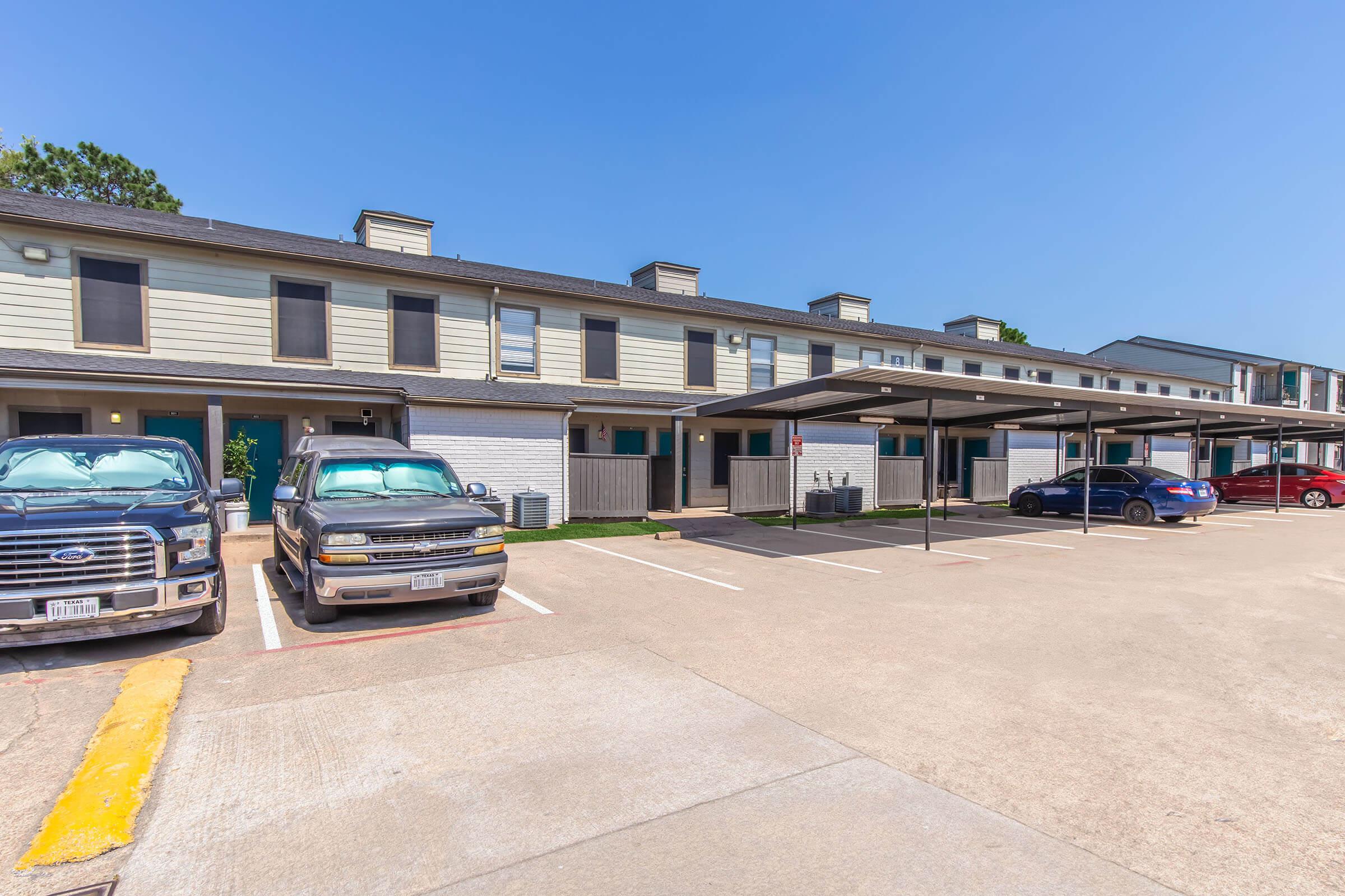 A row of residential townhouses with covered parking spaces in front. Two pickup trucks and a sedan are parked in the lot. The buildings feature a light-colored exterior with several windows and a clear blue sky in the background. Green grass is visible near the parking area.