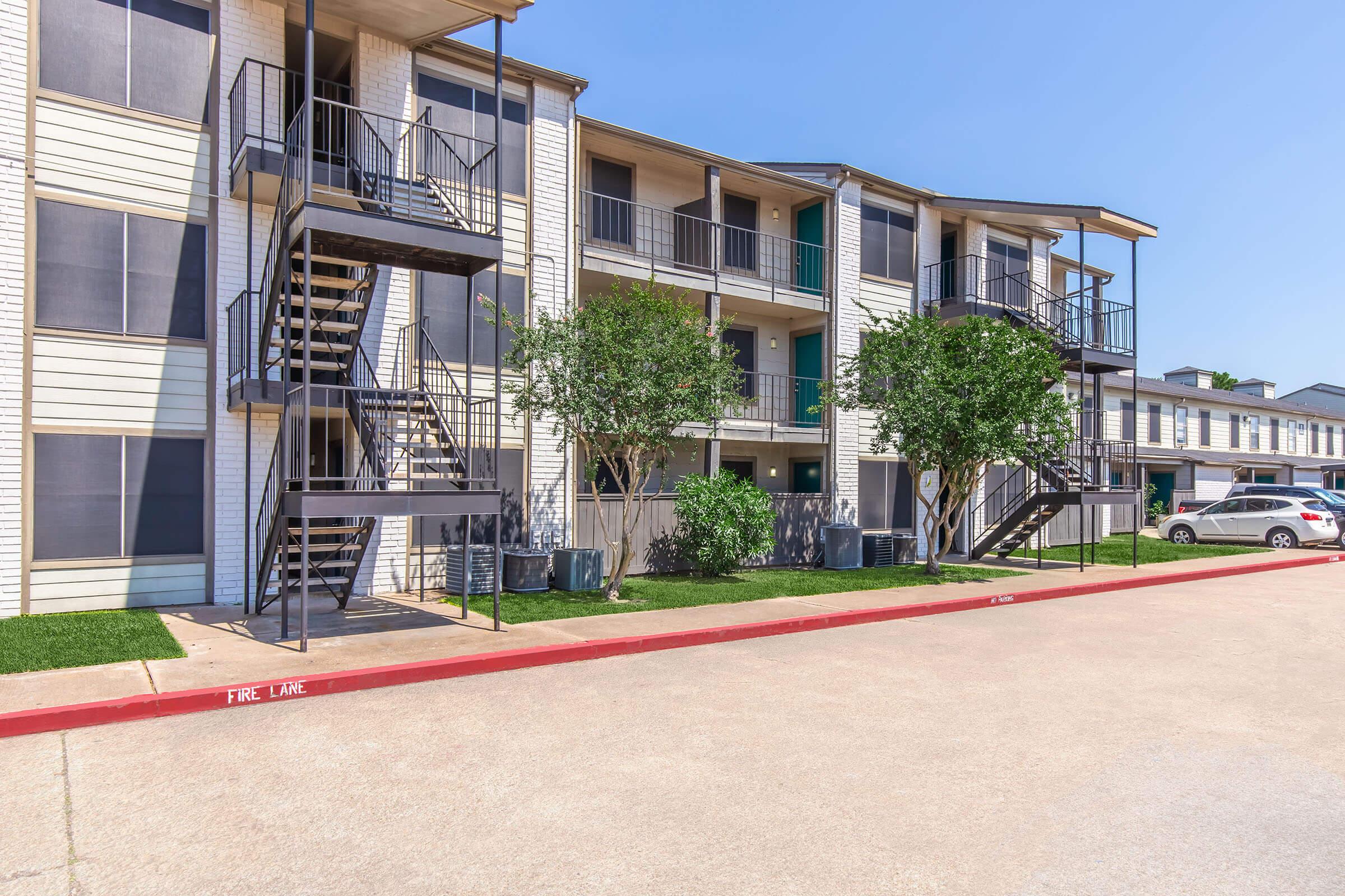 Three-story apartment complex with multiple units, featuring dark metal staircases on the left side and greenery in front. A paved driveway in the foreground shows parked cars, and the backdrop consists of blue sky with a few scattered clouds.