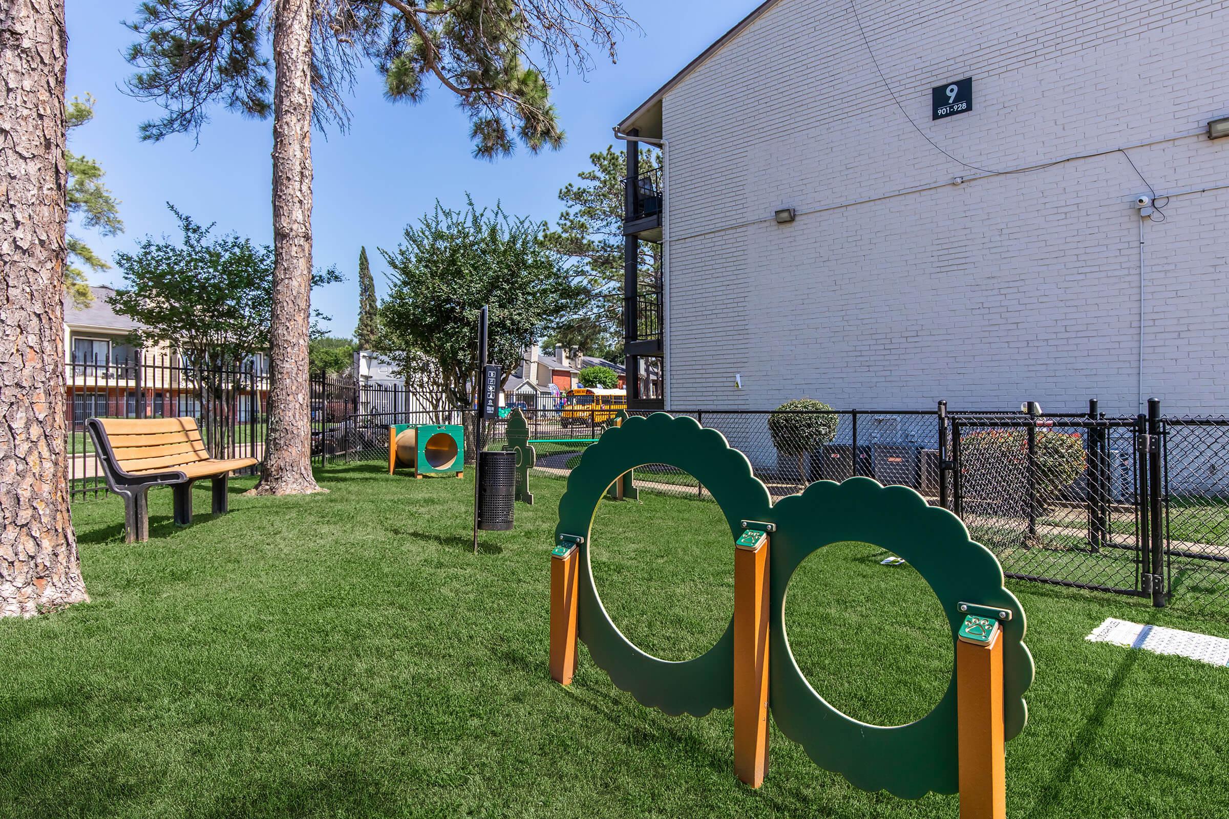 A well-maintained outdoor space featuring dog play equipment, including two large green circular structures for dogs to jump through, surrounded by lush green grass, trees, and a bench. The area is enclosed by a black fence, with an apartment building visible in the background.