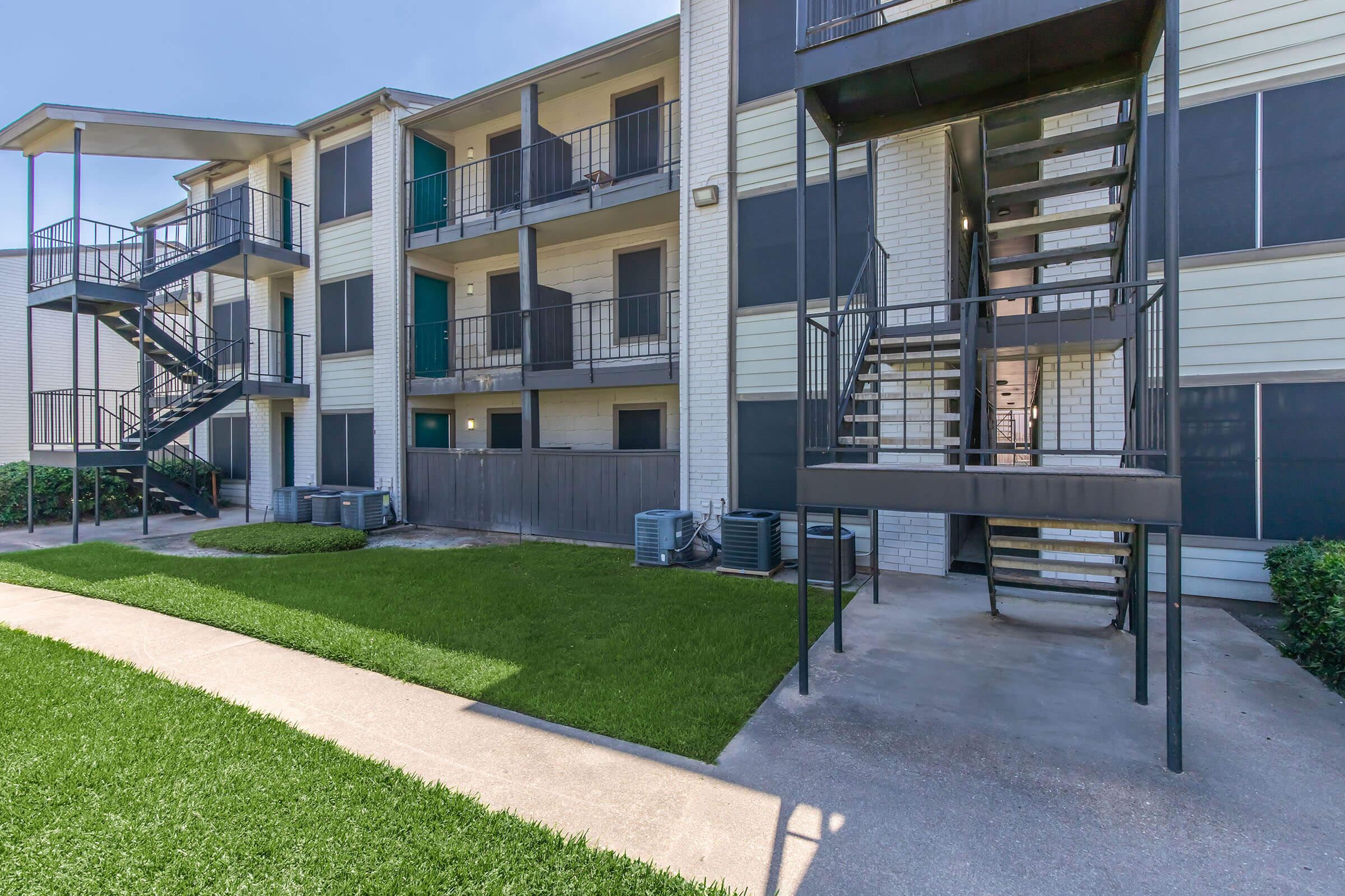 Exterior view of a multi-story apartment building with a staircase and balconies. The building features a combination of dark and light panels, surrounded by well-maintained green grass and a concrete pathway. Air conditioning units are visible on the ground floor.