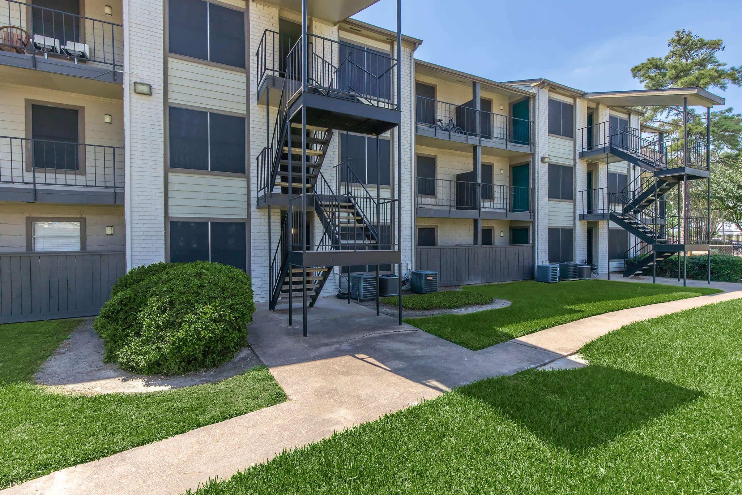 A multi-story residential building with balconies and exterior staircases. The structure features a light-colored facade and is surrounded by green lawns and paved walkways. Air conditioning units are visible at the base of the building. The scene is bathed in sunlight with a clear blue sky above.