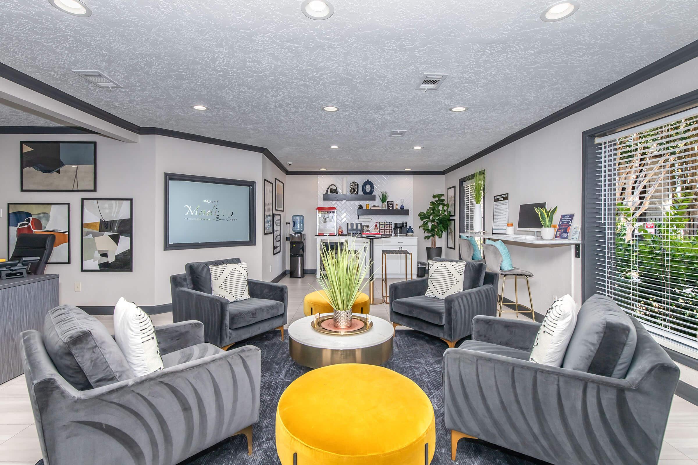 A modern lounge area featuring four plush armchairs and a round yellow coffee table. The space is decorated with contemporary artwork and bright greenery. In the background, there's a reception area with shelves displaying products and a water cooler, all illuminated by ceiling lights.