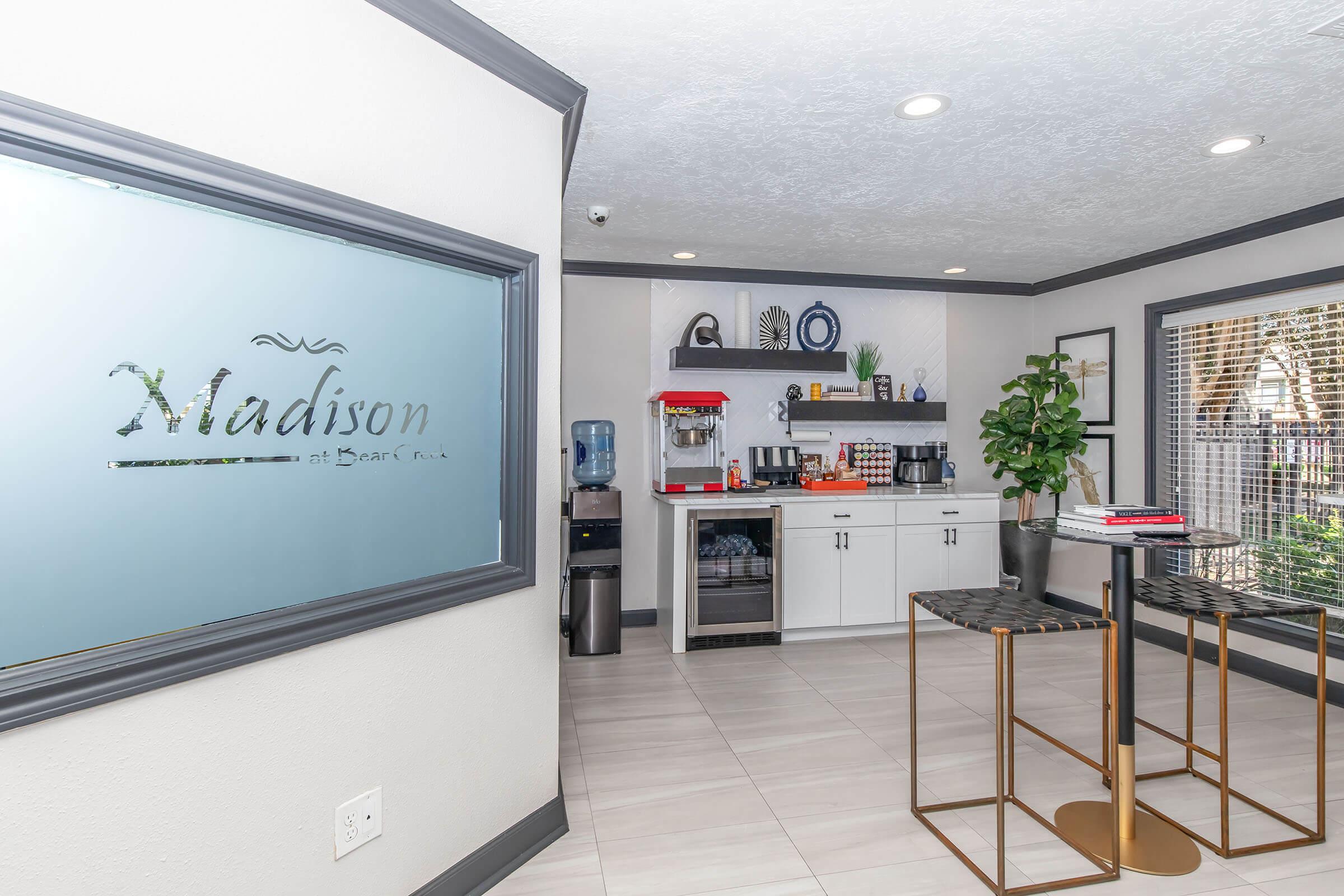 A modern office area featuring a frosted glass sign with "Madison," a kitchenette with a water cooler, coffee maker, and refrigerator, and a small table with gold legs. A potted plant adds a touch of greenery, and large windows allow natural light to brighten the space.