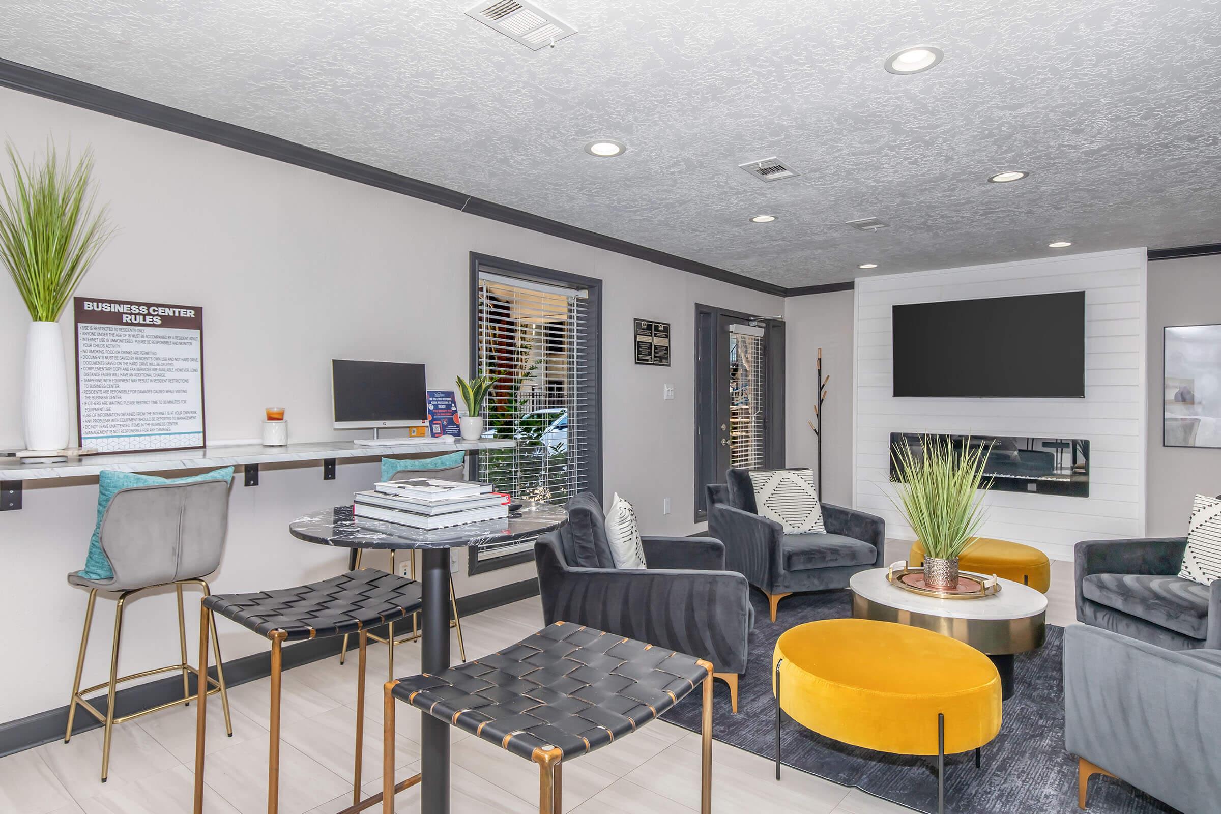 A modern communal lounge area with stylish furniture. It features a circular coffee table with books, two yellow ottomans, and multiple comfortable chairs. A wall-mounted TV is visible, along with a desk and computer station. Large windows provide natural light, and decorative plants enhance the ambiance.