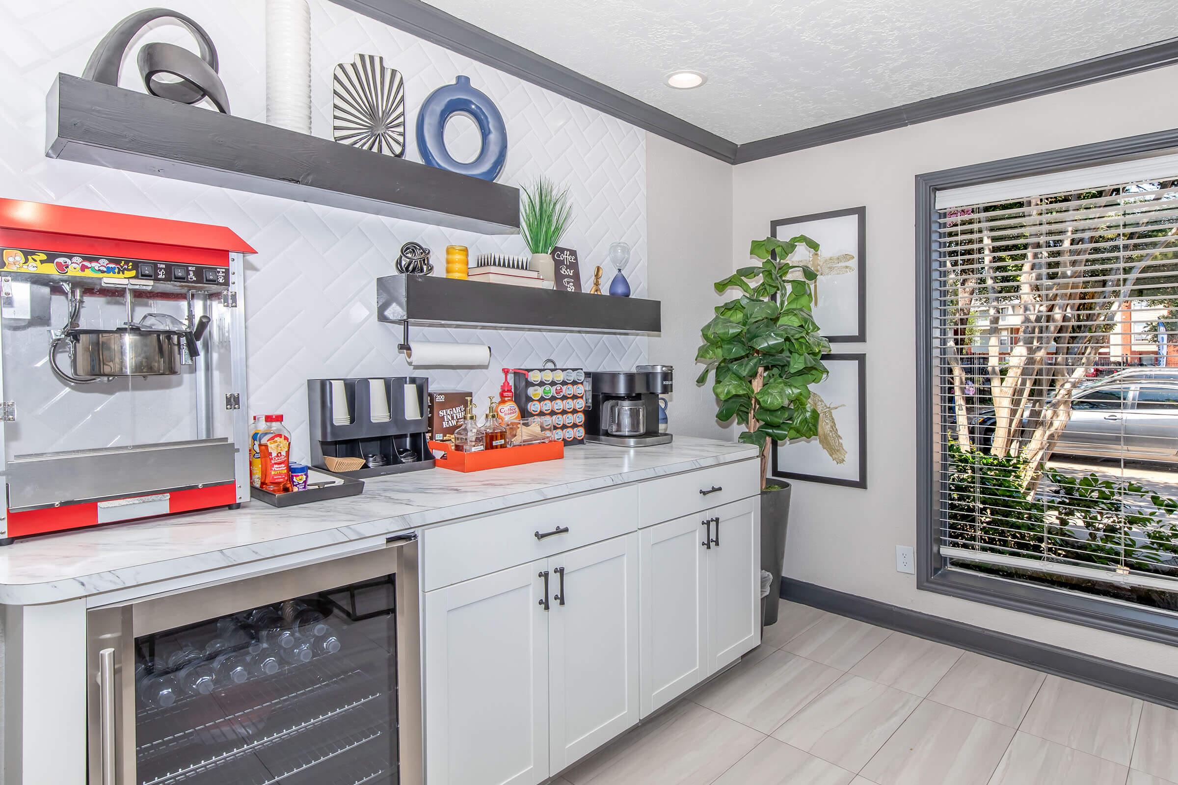 A modern kitchenette featuring a marble countertop with a drink dispenser, coffee maker, and snack display. White cabinetry and open shelves hold decorative items and plants. Large windows let in natural light, illuminating the space with a cozy ambiance.