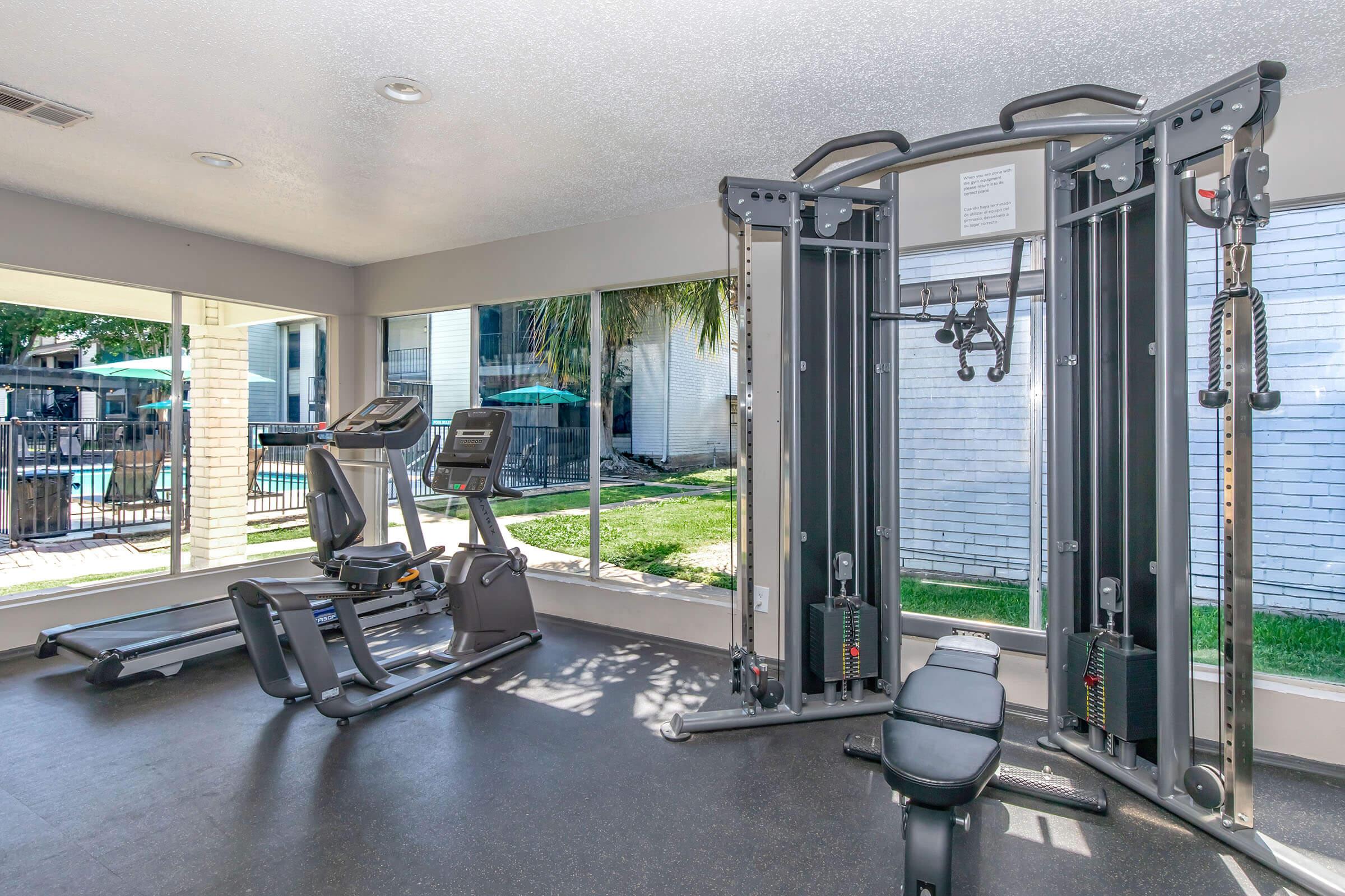 A modern gym interior featuring exercise equipment including a multi-station weight machine, treadmills, and ellipticals. Large windows provide natural light and a view of the outdoor pool area with green grass and patio furniture. The space is clean and well-lit, creating an inviting workout environment.
