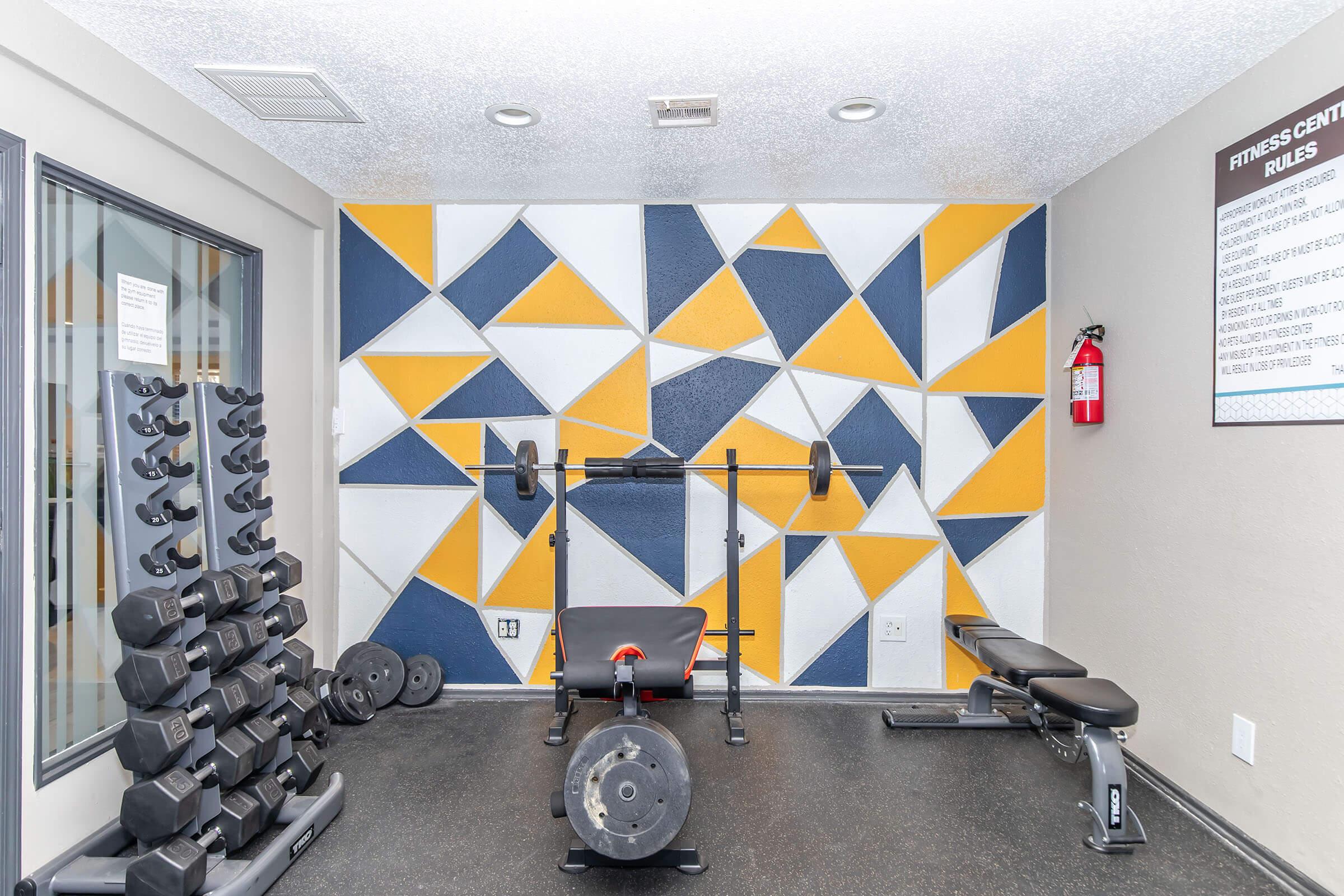 A modern gym space featuring a barbell and bench in the center, surrounded by colorful geometric wall art in yellow, blue, and white. To the left, there is a rack of dumbbells, and the flooring is rubberized, enhancing the fitness environment. A fire extinguisher is visible on the wall.