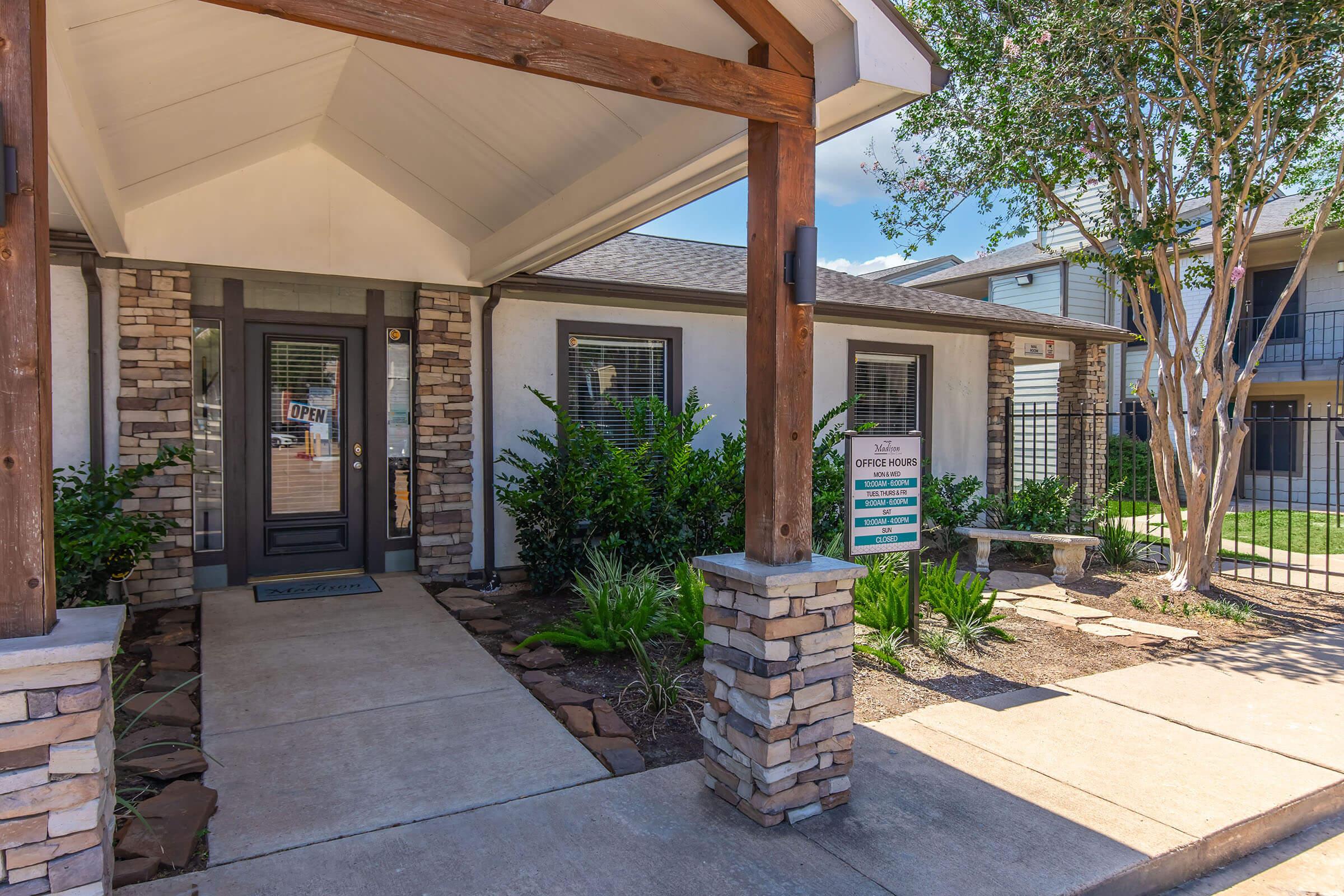 A welcoming entrance to an office building featuring a covered porch with wooden beams and stone pillars. The front door is glass with an "OPEN" sign visible. Lush green plants and landscaping surround the pathway, and a sign for office hours is posted near the entrance.