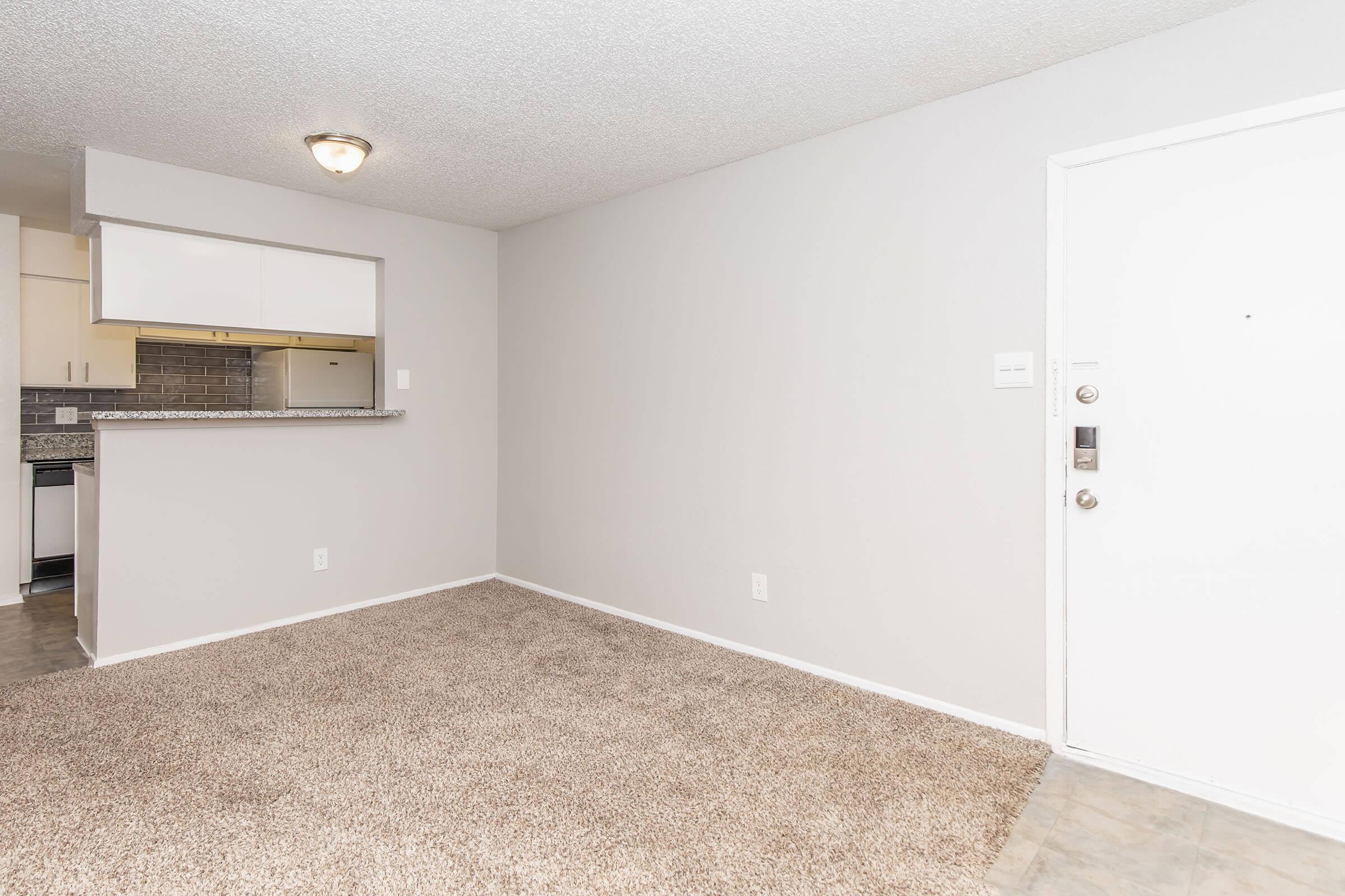 An empty room with beige carpet, a light-colored wall, and a door on the right. There is a small opening leading to a kitchen area in the background with light cabinetry. The ceiling has a light fixture, and the overall atmosphere is neutral and unfurnished.
