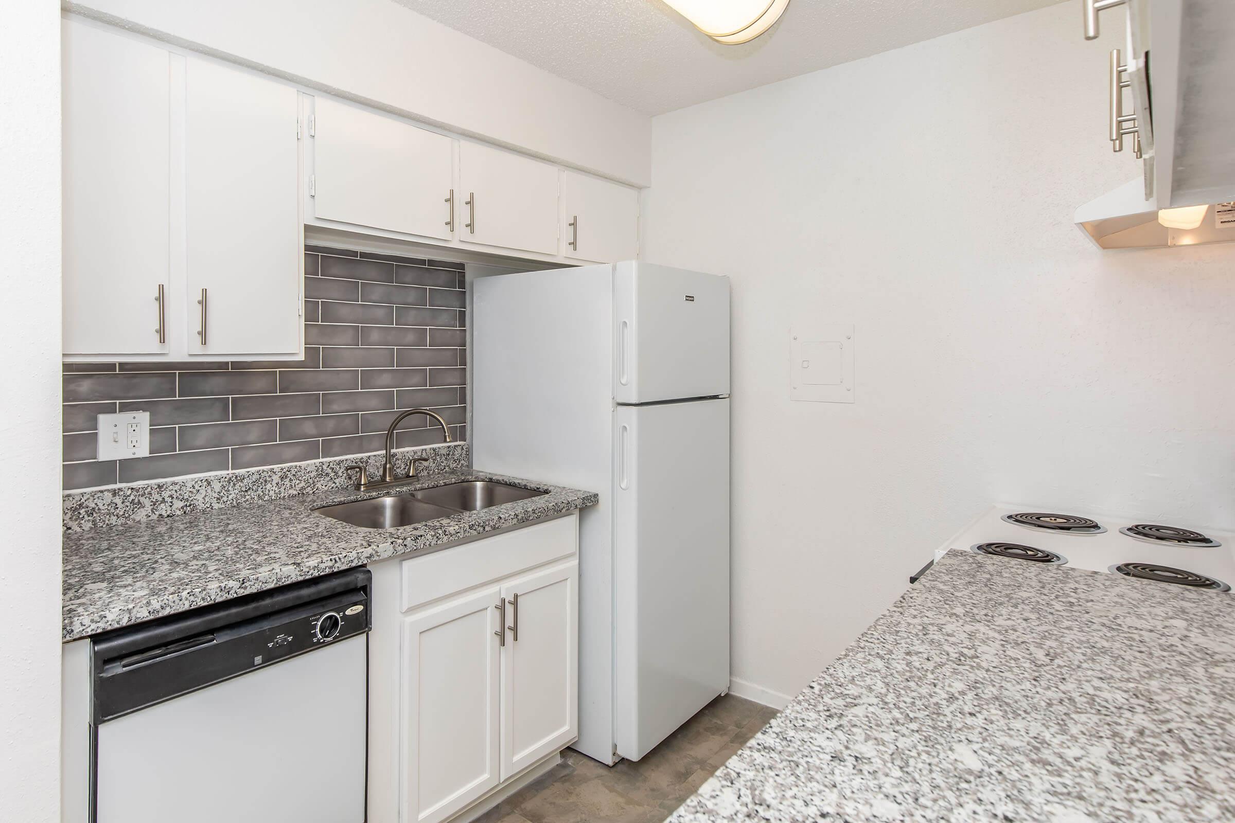 A modern kitchen featuring white cabinets, a double sink, and a granite countertop. There is a gray tiled backsplash, a white refrigerator, and a stove with a cooktop. The overall aesthetic is bright and clean with a minimalist design.